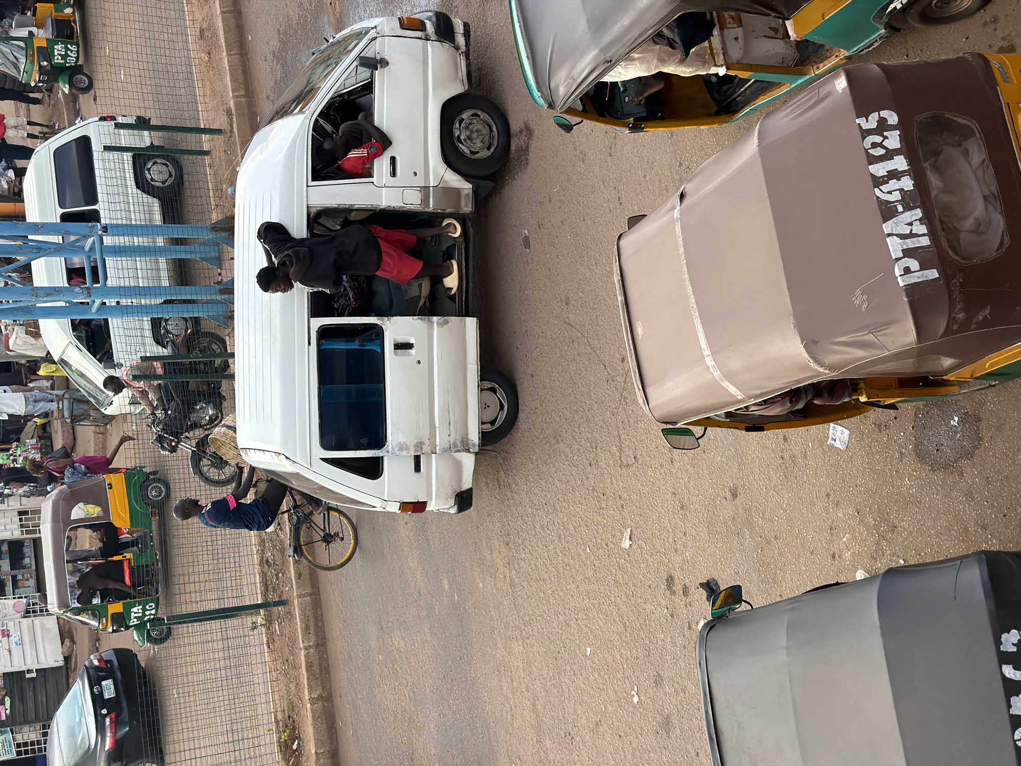 The image depicts a street scene in Bukuru, Nigeria, with a focus on vehicular and pedestrian traffic. In the foreground, a brown and yellow tricycle, identified by the markings "PTA-4125", is parked. Next to it, another tricycle, primarily green and yellow, is also visible. A white passenger van is positioned centrally in the frame, with its side door open, revealing passengers inside. One individual, wearing red shorts and a black hoodie, is standing in the open doorway of the van. Another man in a red shirt leans out from the driver's side window. In the background, a black sedan is parked on the left, and a white commercial van is seen further back. A motorcycle with a rider is also present. Pedestrians are scattered throughout the scene, including a person walking in a pink dress and a man on a bicycle. The environment appears to be a somewhat busy urban street with asphalt pavement and a fence running along the roadside.