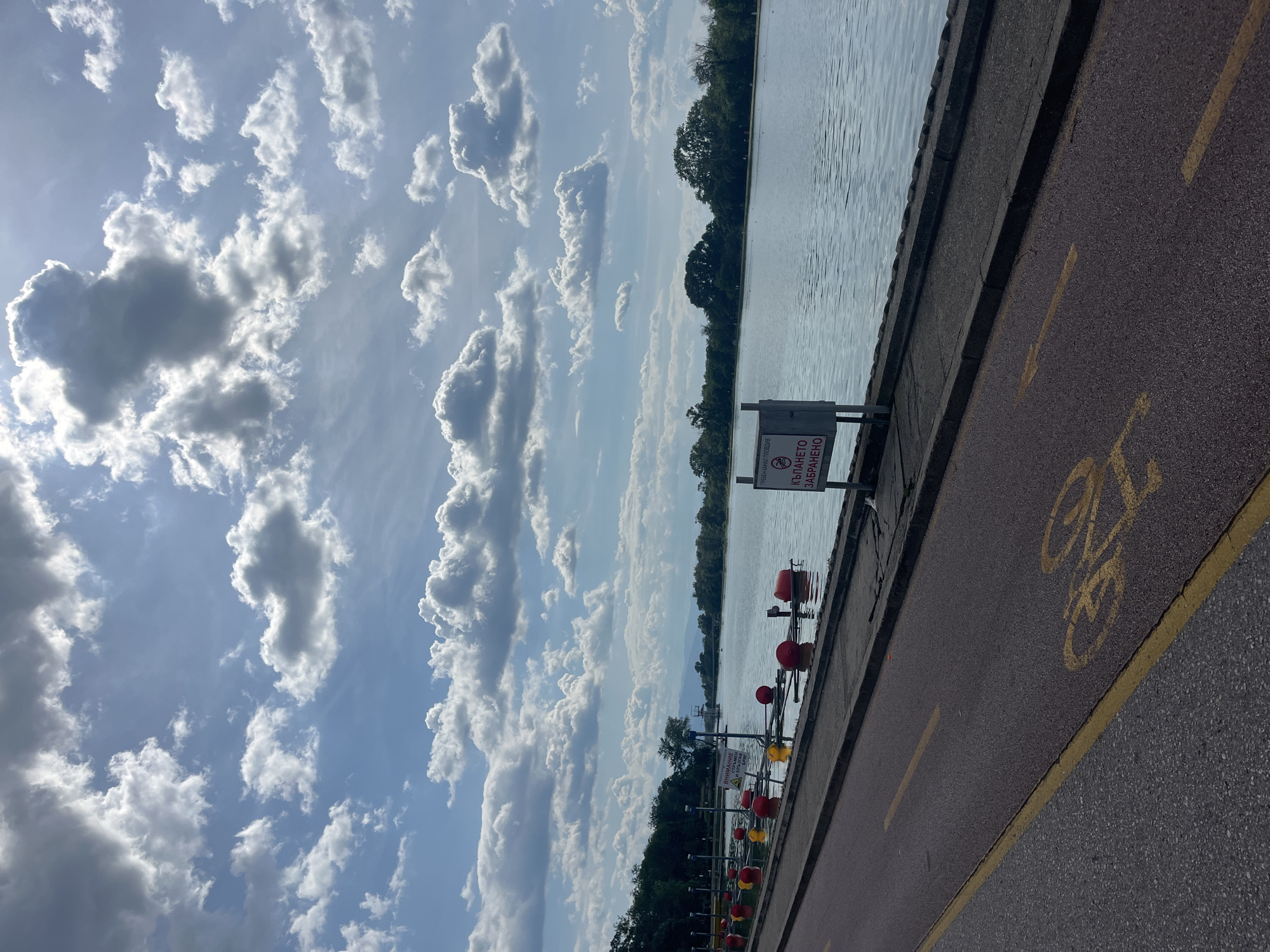 The image depicts a partially cloudy sky above a body of water, possibly a lake or river, next to a paved pathway. A barrier separates the water from the pathway, which features yellow lane markers and a bicycle symbol, indicating it's a bike path. A sign with Cyrillic text, likely a warning or prohibition, is positioned near the water's edge. Red and yellow buoys or markers are visible in the water. Trees line the distant shore.
Based on the Cyrillic text and the scene, the location could be the rowing canal in Plovdiv, Bulgaria, a popular recreational area.