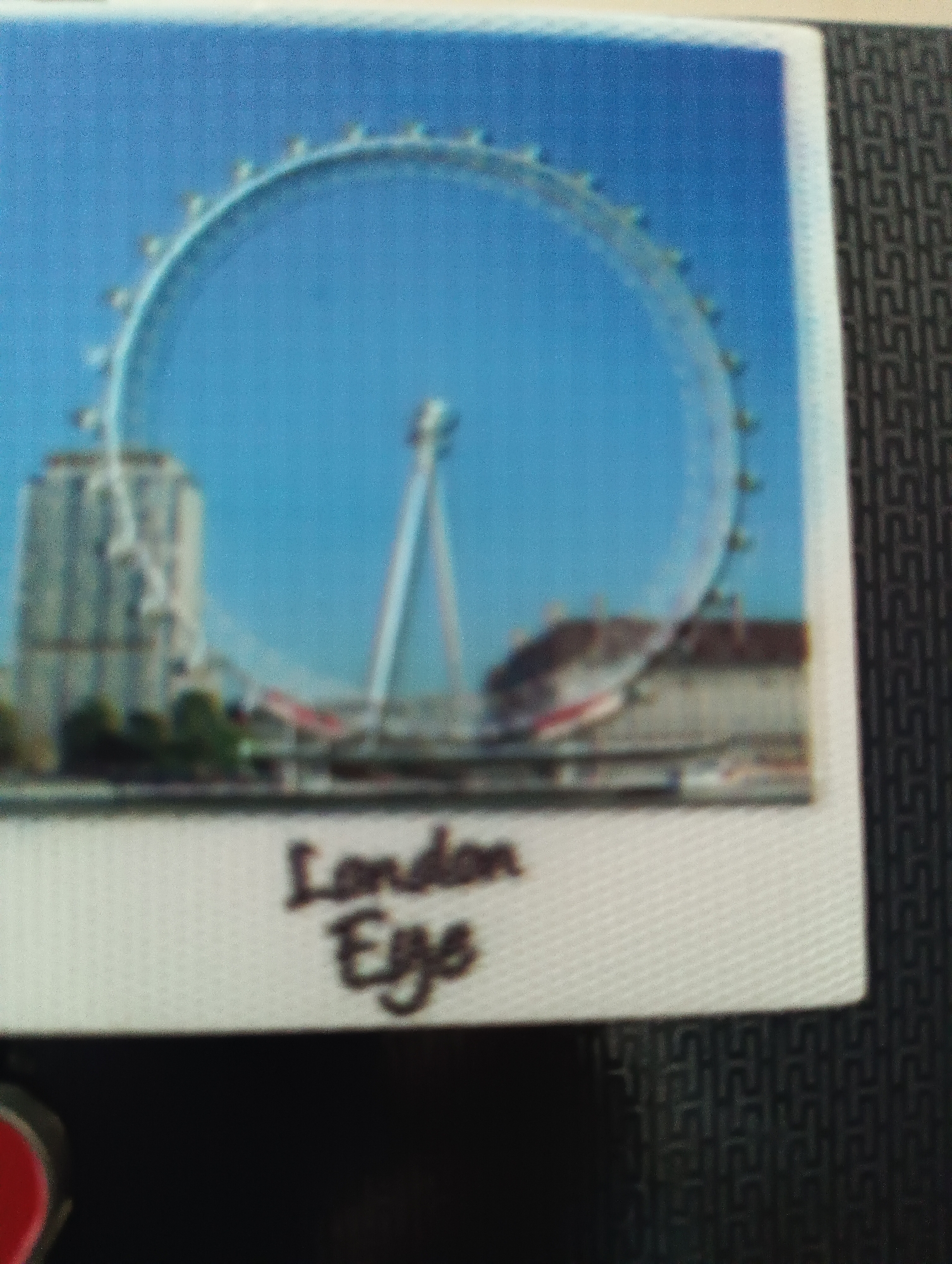 The image is a photograph of a souvenir postcard featuring the London Eye Ferris wheel. The postcard is white with a textured border. The main image depicts the London Eye against a clear blue sky, with buildings and trees in the background. Below the image, handwritten text reads "London Eye". The postcard is placed on a dark, textured surface. The image does not show the city of Karnobat, Bulgaria.