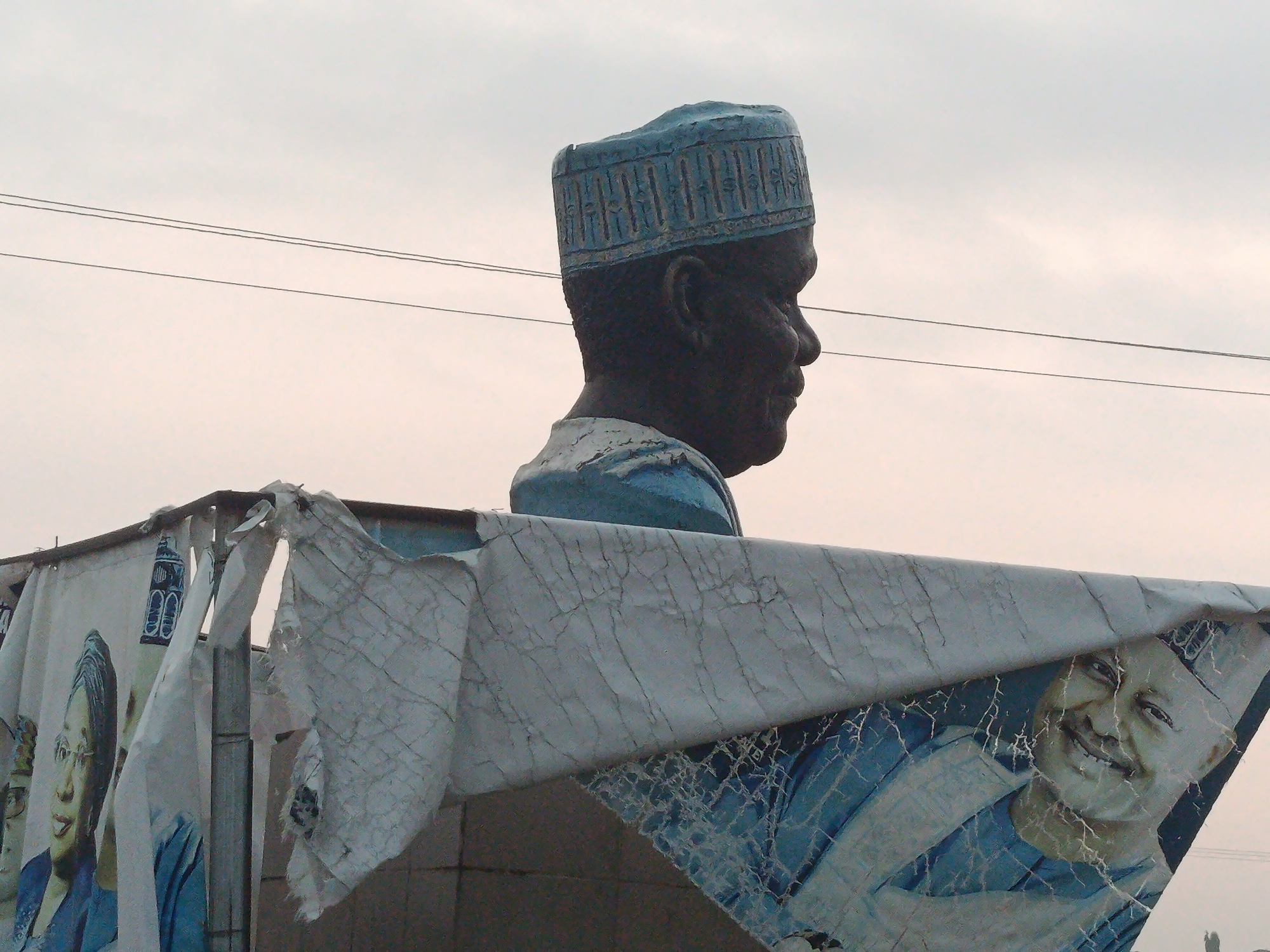 The image shows a large statue of a man wearing a blue patterned hat and blue clothing, seen in profile from the shoulders up. Behind the statue, a torn and weathered banner is visible, featuring images of people. One portion of the banner on the right shows a smiling man in a blue hat and shirt. On the left, a portion of the banner displays a woman wearing glasses and a blue headscarf. The scene appears to be outdoors, with overhead power lines visible against a pale, overcast sky. The statue and banner are likely part of a public display or advertisement in Maigizo, Nigeria, given the context of the provided information. The overall impression is of a weathered and possibly aged display.