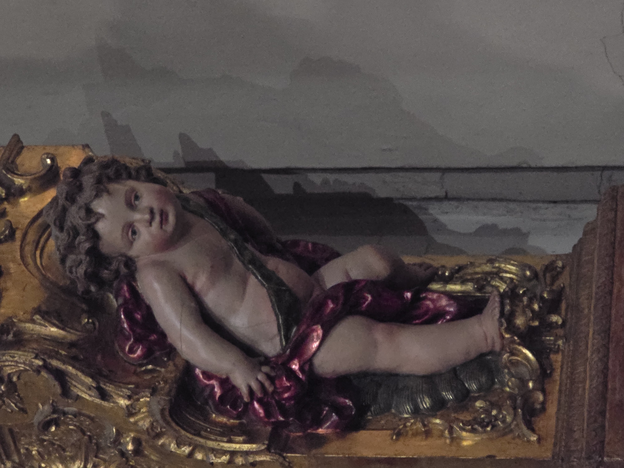 A detailed, forensic, matter-of-fact description of the scene is as follows:
The image shows a close-up, eye-level view of a cherub sculpture, a religious statuary, positioned on the left side of the frame. The sculpture is depicted from the waist down, with its left leg bent at the knee and its foot resting on a decorative gold base. The cherub has fair skin and curly brown hair. It is draped in a deep red fabric with a green sash. The sculpture is ornate, with a gilded frame featuring swirling, elaborate designs. The background consists of a neutral-toned wall with subtle shadows cast by the sculpture and its surrounding structure. The lighting appears to be ambient, with some directional light highlighting the sculpture's features and casting shadows. The overall color palette is warm, dominated by gold, red, and the pale tones of the cherub's skin.