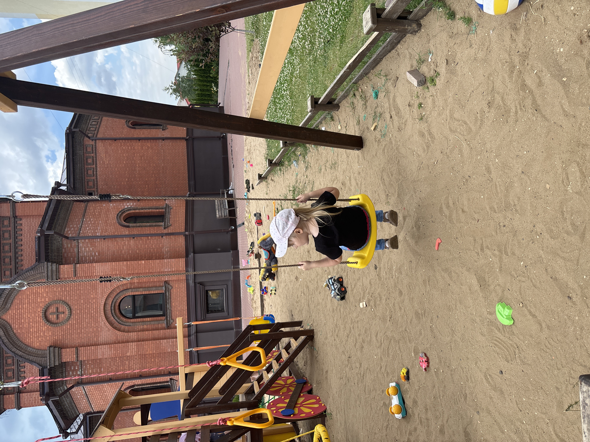 This is a medium-angle shot of a child on a swing set, situated in a sandy playground. The child, wearing a white cap and a black shirt, is actively engaged in swinging on a yellow seat. The playground is littered with various toys, including toy cars, a green sand mold, and a pink toy. A portion of a wooden playground structure with yellow handrails is visible in the foreground. In the background, a large brick building with arched windows and architectural details stands prominently. The sky above is partly cloudy, suggesting fair weather. The scene appears to be set during the daytime. The overall atmosphere is one of children's play and recreation. The location is identified as Saratov, Russia. There is no visible text in the image.