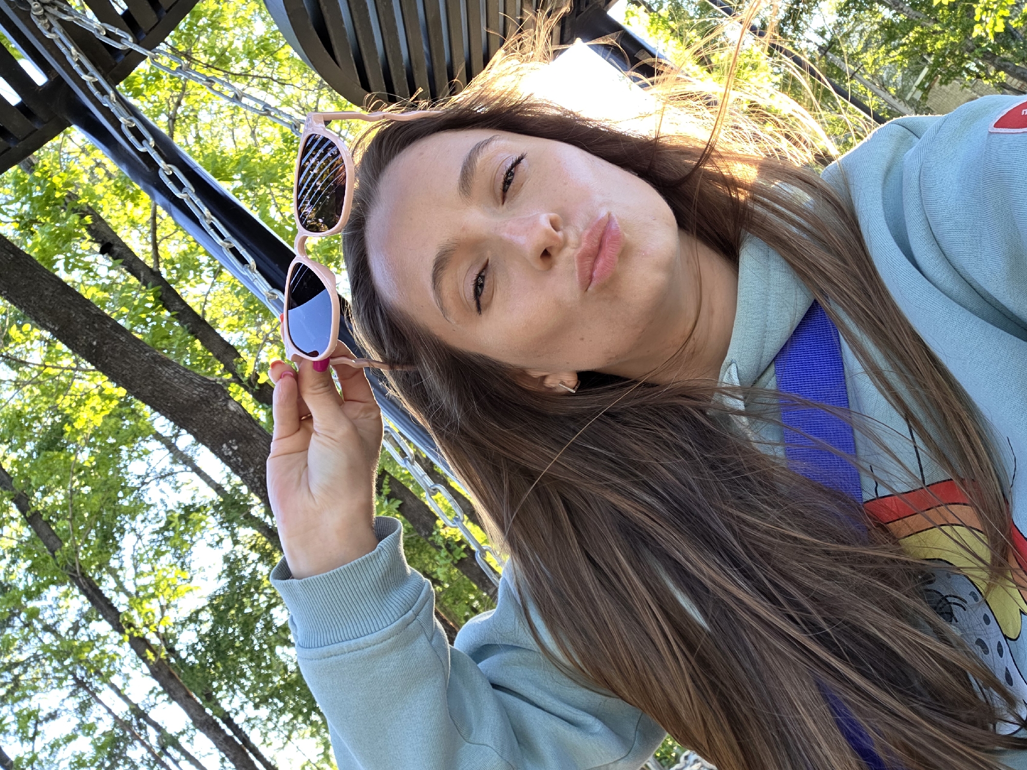 A person with long brown hair is taking a selfie. They are holding pink-framed sunglasses in their left hand. The person is wearing a light blue sweatshirt with a graphic design visible on the right side. A blue strap is also visible. The background shows trees and a metal structure with chains, possibly a swing or climbing frame. The location is not definitively identifiable as Birobidzhan, Russia, based solely on the image content.