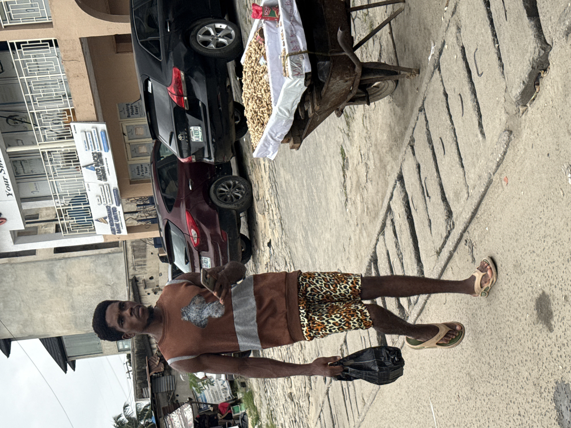 A Black male walks on a concrete sidewalk on a street in Ado, Nigeria, holding a black plastic bag in his left hand and a smartphone in his right. He wears a sleeveless brown shirt with a gray stripe across the chest and leopard-print shorts. He is also wearing flip-flops. Beside him on the right, a wheelbarrow is filled with peanuts. In the background, cars are parked along the street. The street is paved with concrete slabs and there are buildings in the background.