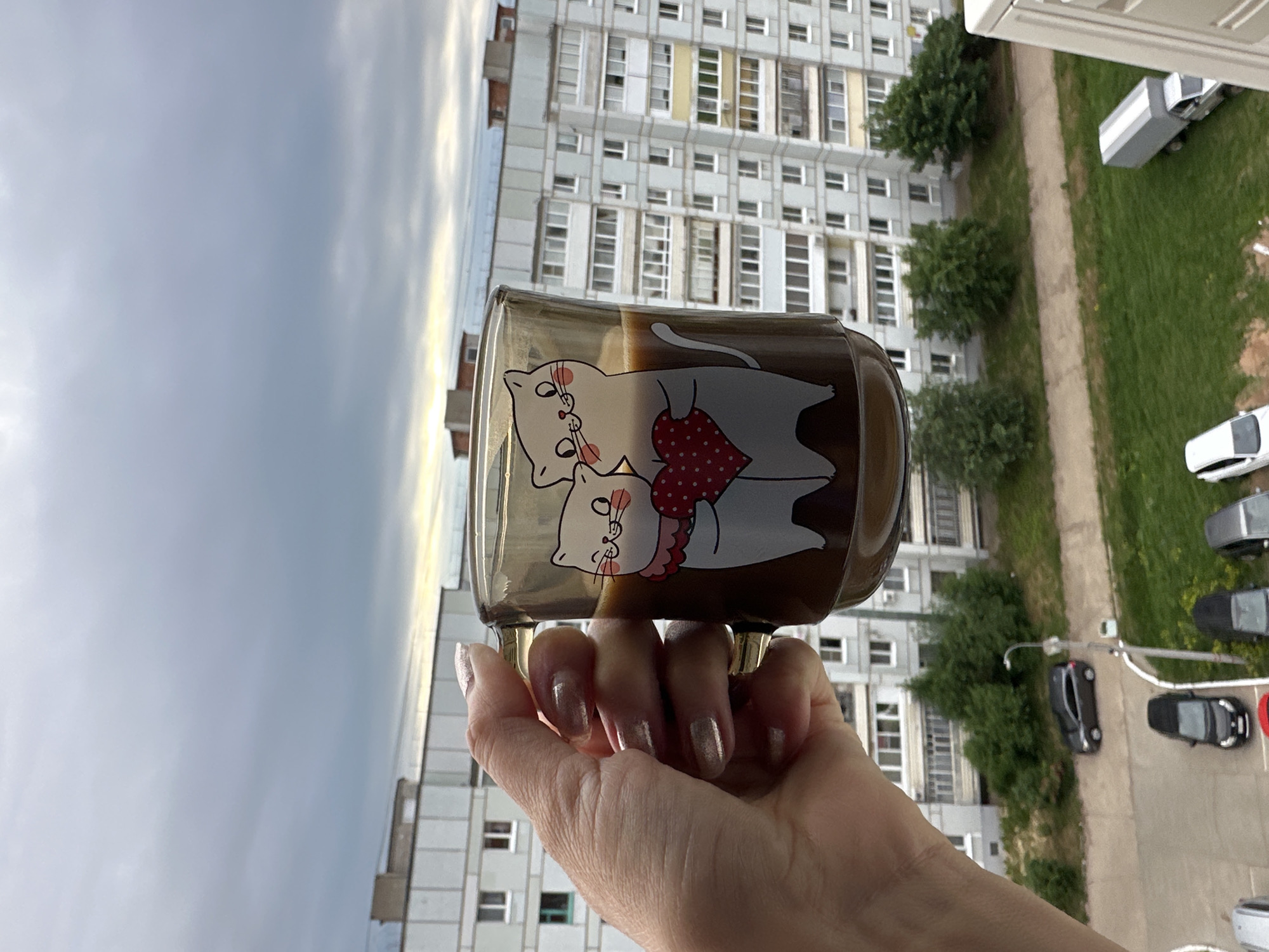 A hand with silver glitter nail polish holds a transparent mug filled with coffee. The mug features a cartoon illustration of two white cats embracing, with one holding a red heart with white polka dots. In the background, a multi-story apartment building with numerous windows dominates the scene. Trees and parked cars are visible in front of the building, suggesting an urban residential area. The sky is overcast with gray clouds. The image appears to be taken from a balcony or window in the city of Chaikovsky, Russia, given the architectural style of the building.