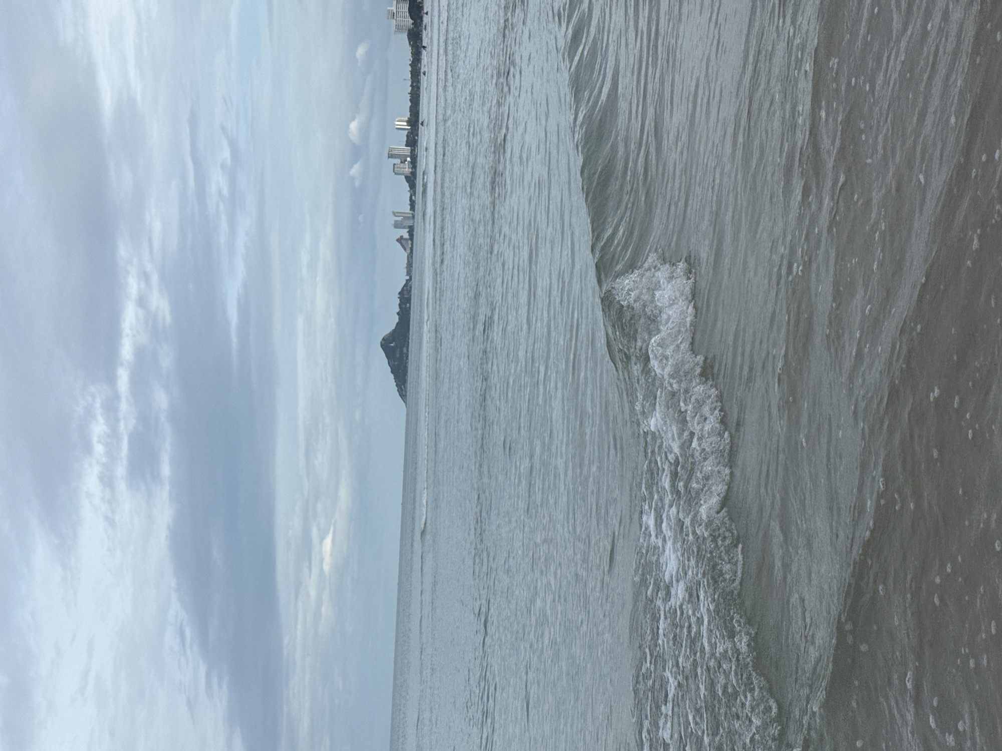 A wide shot captures a beach scene in Hua Hin City Municipality, Thailand, under a cloudy sky. The foreground is dominated by the calm, gray ocean with gentle waves lapping at the shore. In the mid-ground, a line of buildings, some appearing to be hotels or condominiums, stretches along the coastline. To the left of the buildings, a distinctive conical mountain juts out from the land. Further to the right, a few scattered figures are visible on the beach or in the water, with some appearing to be engaged in water sports like jet skiing. The overall atmosphere is serene, with the overcast conditions contributing to a soft, diffused light.