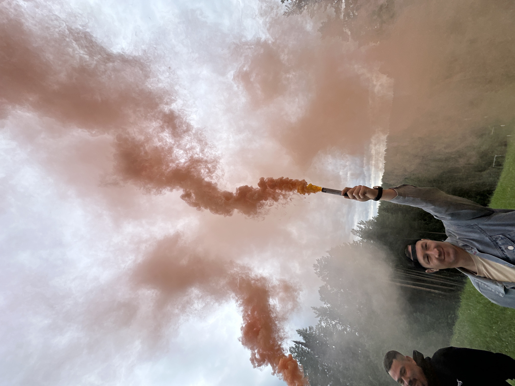 Two men stand in a grassy field in Прикамский, Russia. One man, wearing a denim jacket and black headband, holds a lit orange smoke grenade above his head. The other man stands near the bottom of the frame wearing a black hoodie. Orange smoke fills the sky and field in front of them. The sky is overcast. Trees line the background.