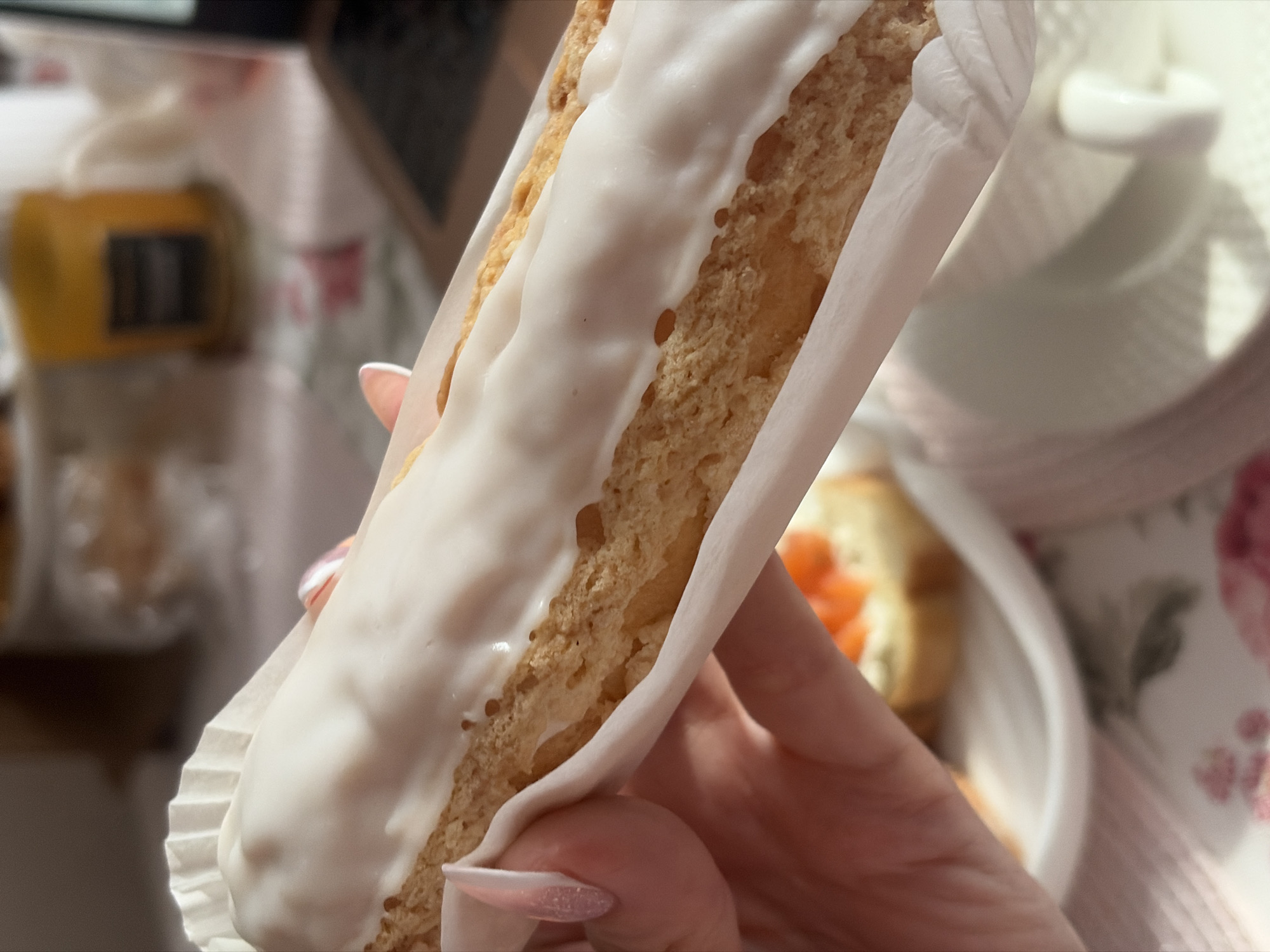 The image features a long, rectangular pastry, possibly an éclair, held in a white paper wrapper. It is topped with a thick, smooth layer of white icing. The pastry has a golden-brown, textured surface beneath the icing. A hand with manicured nails, painted a light pink with glitter, is holding the pastry.
In the blurred background, there is a table with a floral-patterned tablecloth. A white dish containing other food items, some with an orange hue, is visible. A yellow container and other unidentifiable objects are also present in the background.
The location is likely a café or restaurant in Saint Petersburg, Russia, judging by the European-style pastry and the general aesthetic of the setting.