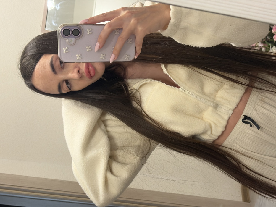 The image is a mirror selfie showing a young woman indoors. She has long, straight, dark brown hair that flows over her shoulders and chest. She is wearing a cream or off-white outfit, consisting of a cropped, long-sleeved jacket made of a soft, fuzzy, fleece-like material, featuring a white zipper down the front. Matching ribbed pants or shorts are partially visible on her lower right side, with a small dark emblem or design near the waist.
Her left hand is holding a smartphone, positioned in the upper central part of the image, capturing her reflection. Her fingernails are long, almond-shaped, and painted a light pink. The smartphone case is a distinct lavender or light purple color and is decorated with multiple small, white, flower-shaped embellishments.
The woman's face is visible, turned slightly towards the camera with her dark eyes looking out from under well-defined eyebrows. Her lips are full and slightly parted in a soft pout.
The background reflected in the mirror appears to be a plain, light-colored wall. In the upper right corner of the reflected space, there is a soft, out-of-focus hint of pink, which could be artificial flowers or a decorative element. The entire image is oriented diagonally, indicating it was taken at an angle or has been rotated. The lighting in the scene is soft and even, suggesting an indoor environment without harsh shadows.