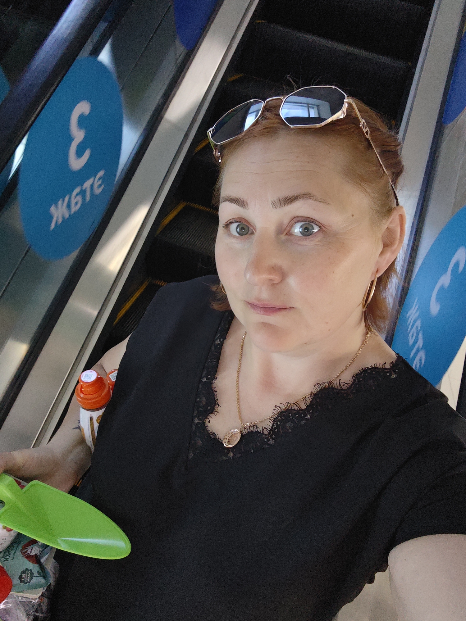 The image is a selfie of a middle-aged woman on an escalator, seemingly inside a shopping mall or public building. She has red hair, fair skin, and is wearing sunglasses perched on her head, a black top with lace detailing, and gold hoop earrings. She has a gold necklace with a pendant.
She is holding a green plastic toy shovel and an orange and white water bottle.
The escalator has blue signage with white Cyrillic letters. The location is the city of Chaikovsky, Russia. The signage contains Russian letters.