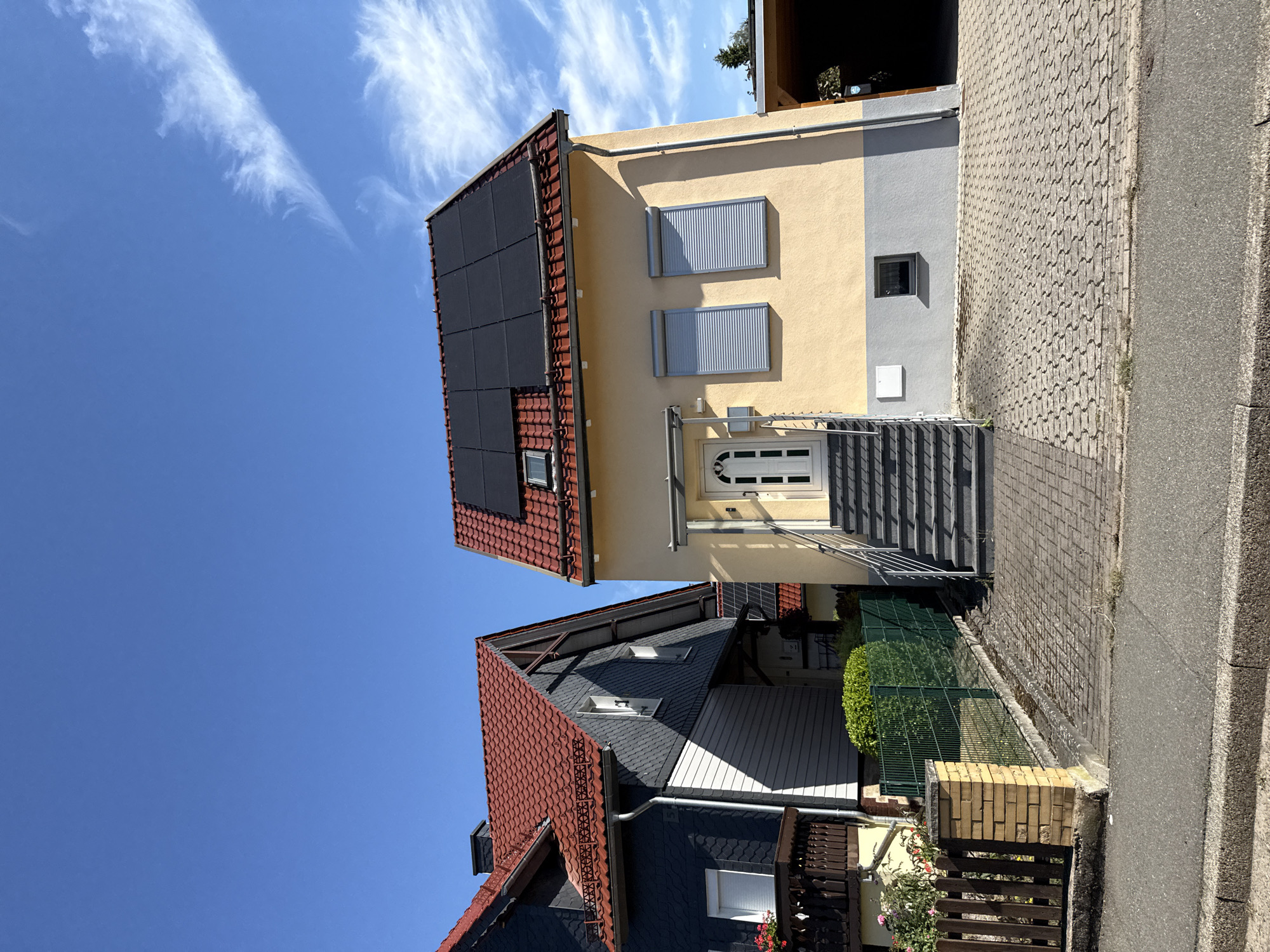 The image shows two adjacent houses on a sunny day in Bad Sachsa, Germany. The house on the right is a light yellow, two-story building with a red tiled roof covered in black solar panels. It has a white front door with a glass pane, accessed by a set of grey metal stairs. Two windows on the upper floor are covered by grey rolling shutters. The house on the left is darker, with grey slate cladding on the upper portion and light grey siding on the lower portion, also with a red tiled roof. A wooden fence with flower boxes and a brick pillar separates the properties from a paved driveway and sidewalk. The sky is bright blue with some wispy white clouds.