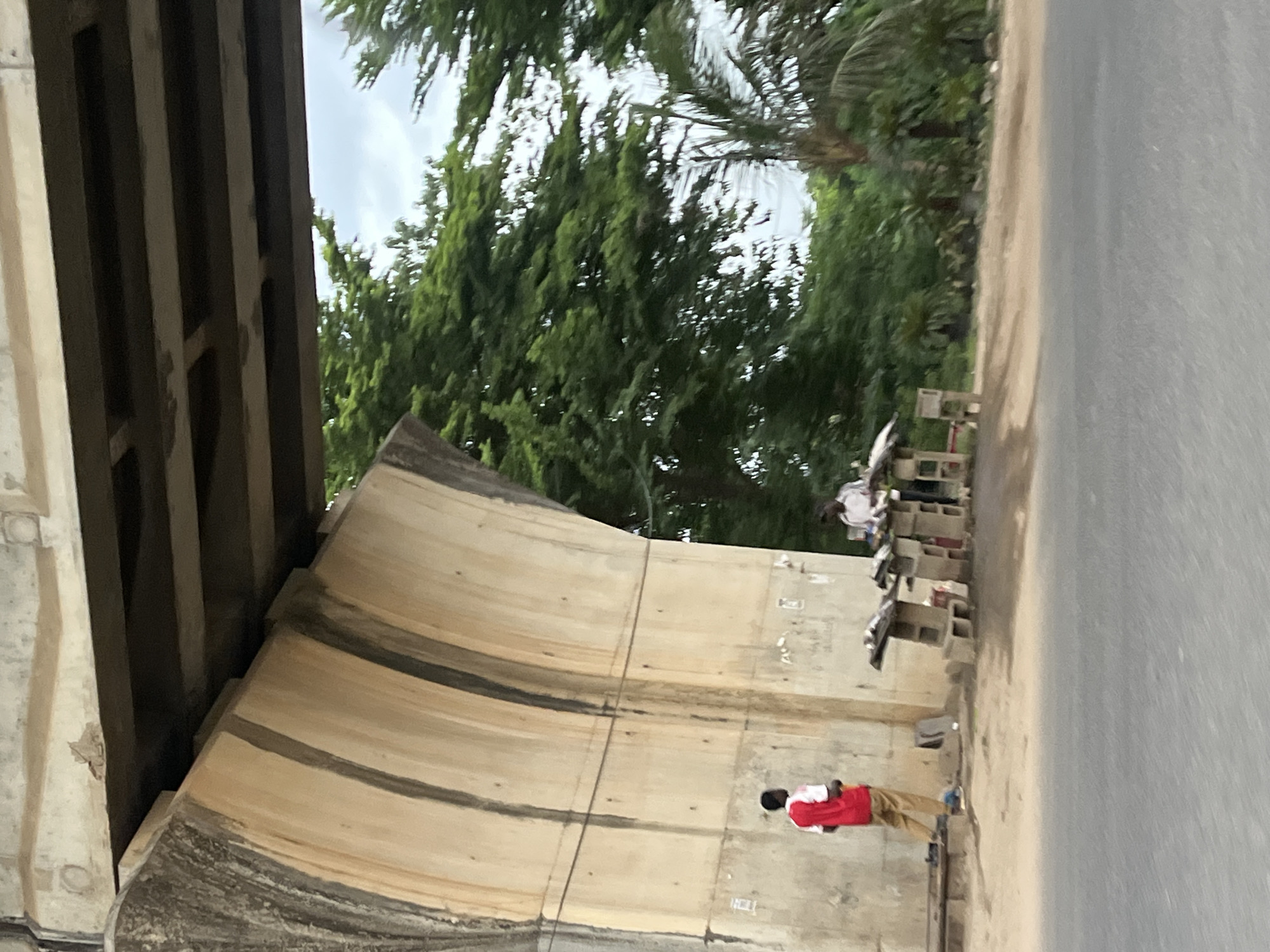 The image depicts a scene under an overpass or bridge, likely in Karmajiji, Nigeria. A person wearing a red and tan outfit stands on the sidewalk. An informal roadside market is set up near the overpass, with what appears to be cinder blocks, merchandise, and other individuals present. The road surface is visible. Trees and vegetation are visible beyond the roadway. The architecture of the overpass features a curved, segmented design with visible seams or markings.