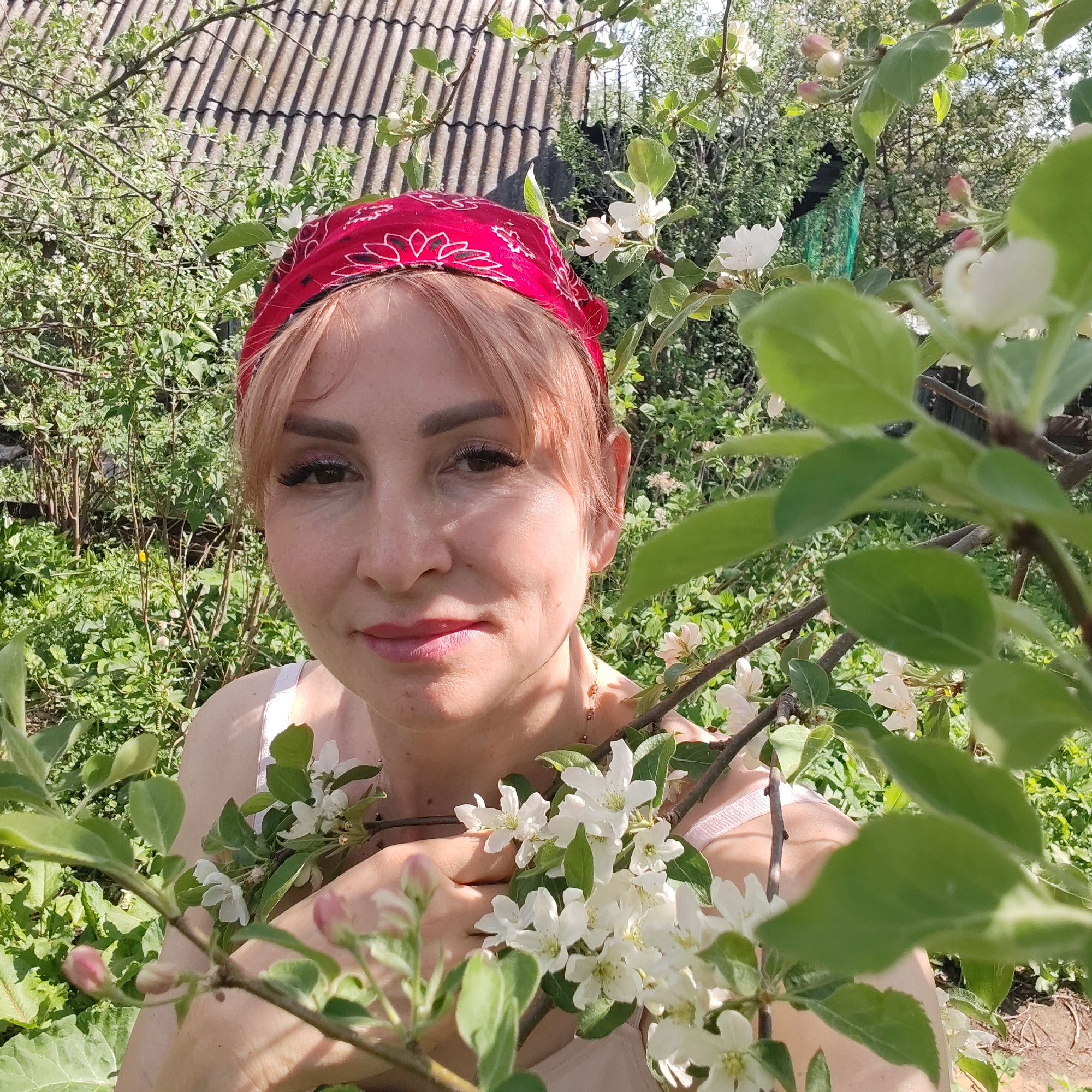 A woman with light reddish-blonde hair, wearing a red patterned bandana, is the primary subject in the image. She is surrounded by green foliage and branches with white flowers, possibly in a garden. Her expression is soft and smiling. She is wearing a light-colored top with thin straps.
In the background, behind the foliage, there is a partially visible roof of a building with gray tiles. The setting is outdoors, with various plants and greenery filling the frame. The image captures a moment in a natural environment, with the focus on the woman amid the blossoming flowers.
The location in the city of Chaikovsky, Russia is not discernible from the image alone.