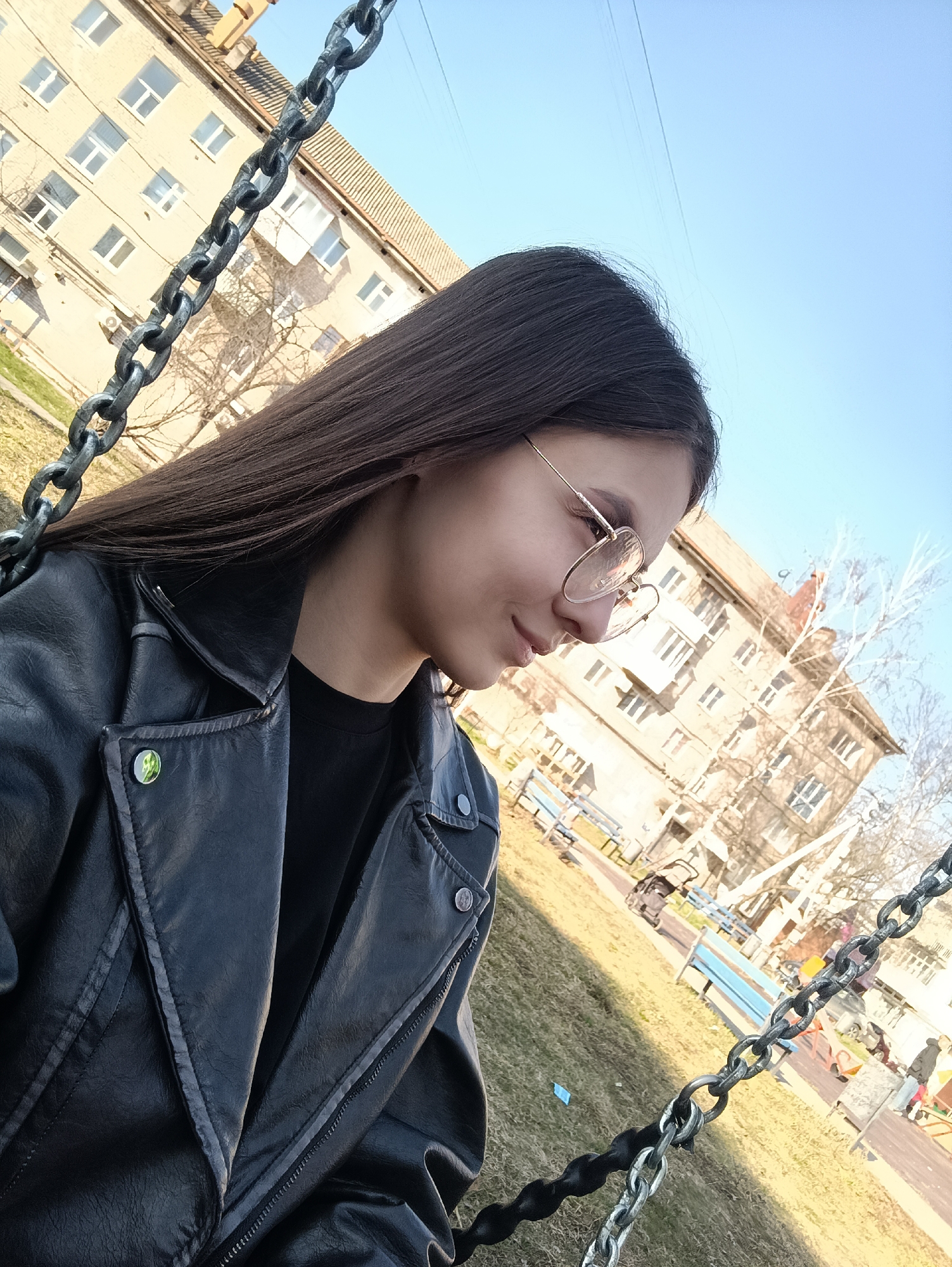 The image shows a young woman in a black leather jacket and glasses, sitting on a swing. The location appears to be a playground in a city, with apartment buildings in the background. There are chains from the swing in the foreground, and trees and benches are visible in the background. The sky is blue and the scene is well-lit, suggesting it is daytime.