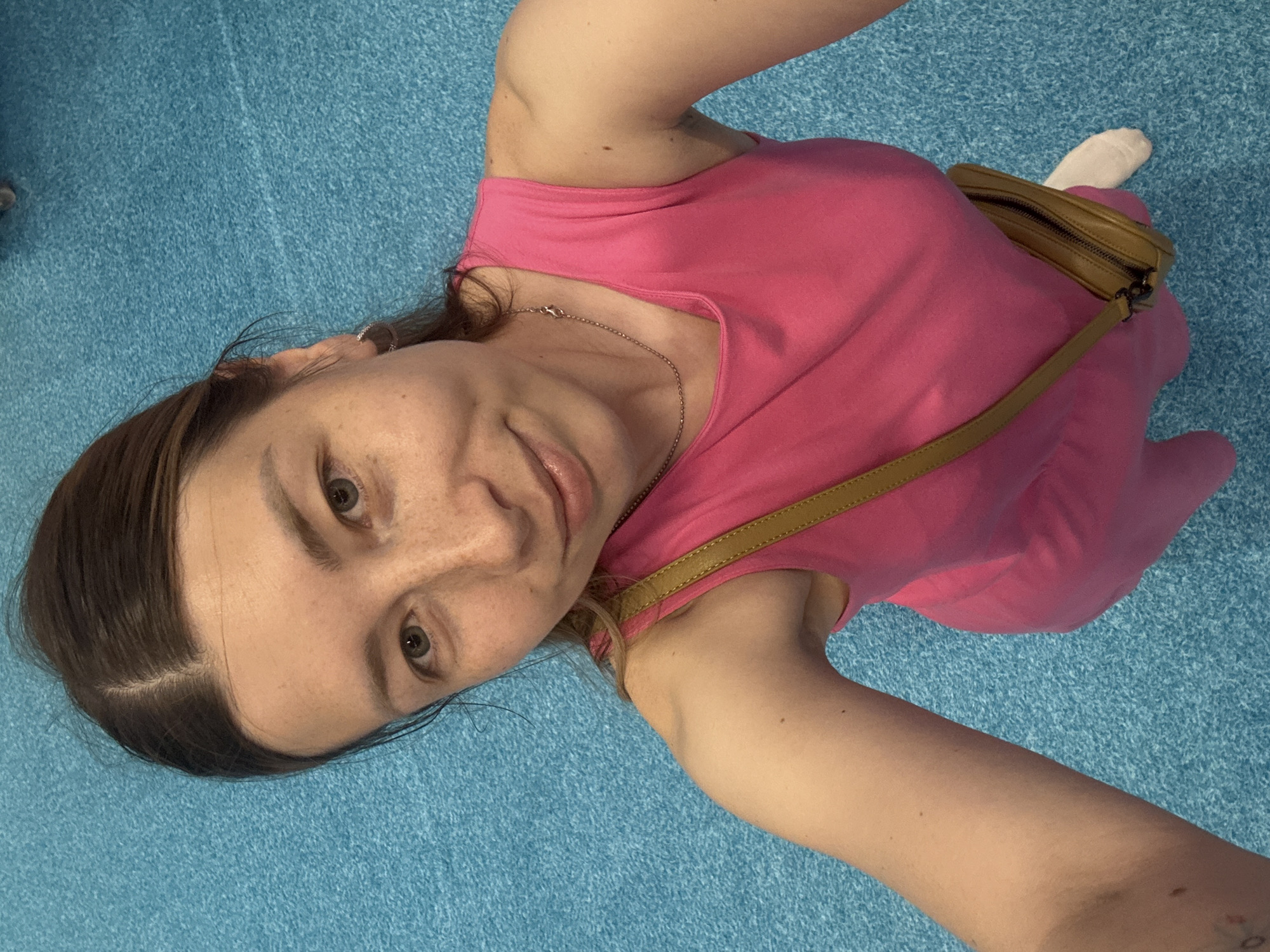 A woman wearing a pink dress and a light brown shoulder bag poses for a selfie. She is lying on a blue surface, presumably a carpet or rug. She is wearing a silver chain necklace. The location is unknown; there is no indication it is in Перовское сельское поселение, Ukraine.