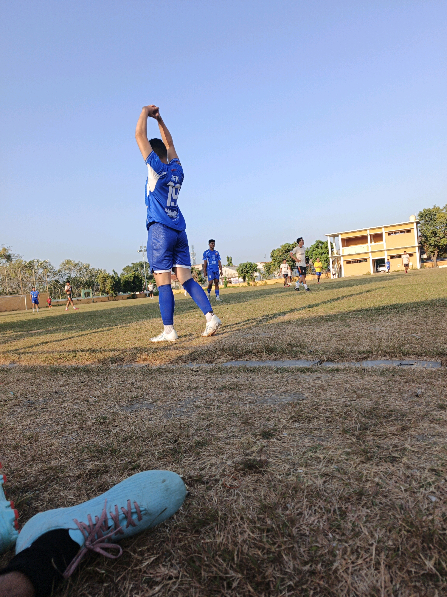 A group of young men in soccer uniforms are practicing on a dry grass field with a building in the background. The man in the foreground, wearing a blue jersey with the number 19, is stretching with his arms above his head. Other players are scattered across the field, some with their backs to the camera. The sky is clear and blue, indicating it is daytime. The scene appears to be a sports training session at a school or local park in Mojokerto, Indonesia.