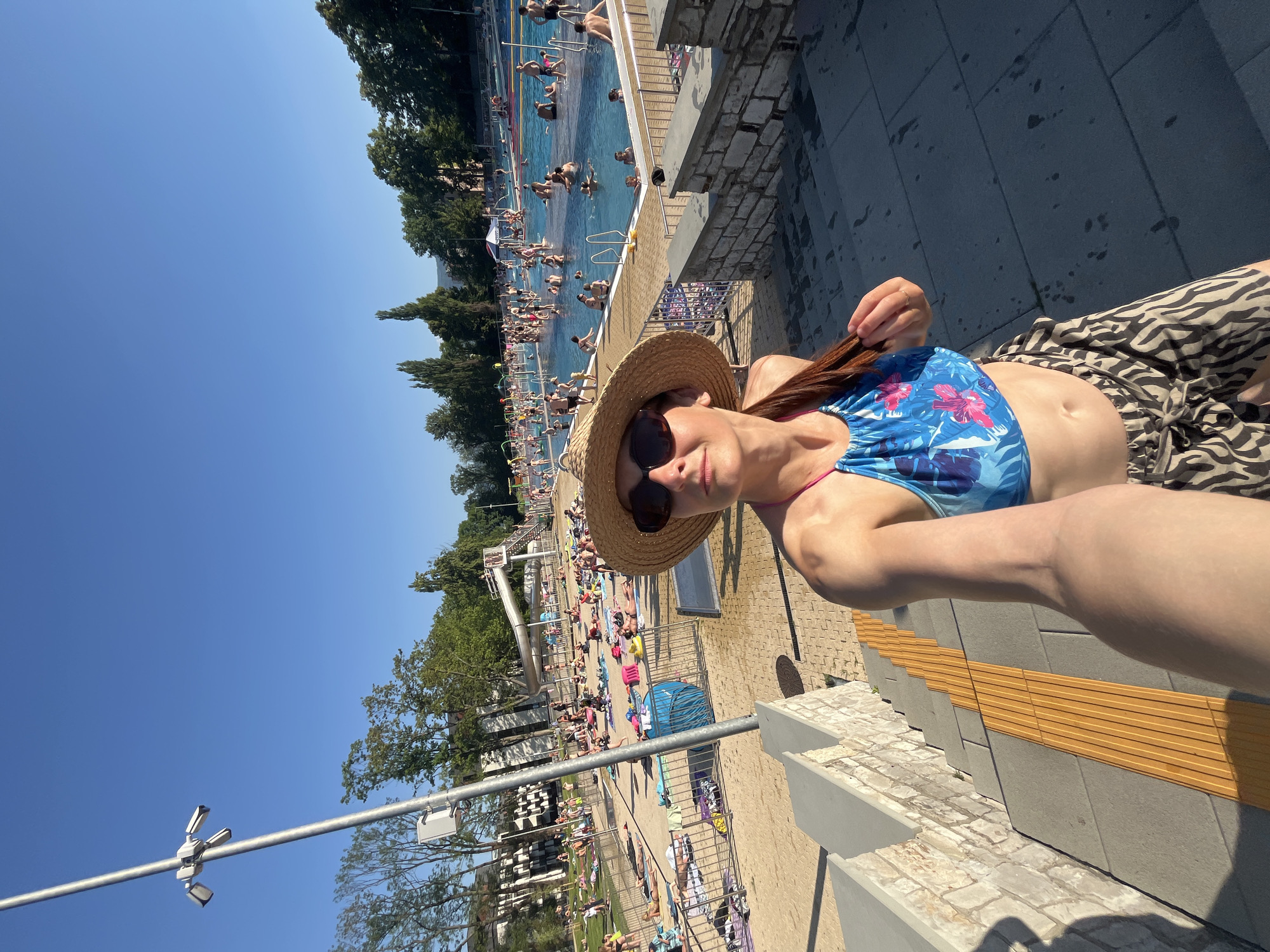 The image depicts a busy outdoor swimming pool on a sunny day. A woman in a blue and pink floral bikini and a straw hat is in the foreground, taking a selfie. Behind her, the large pool is filled with many people swimming and relaxing. Surrounding the pool is a sandy area with lounge chairs and umbrellas, where more people are sunbathing. Trees and buildings are visible in the background, suggesting a developed recreational area. The bright sunlight indicates it is daytime, likely midday, and the clear blue sky suggests fair weather. The overall atmosphere appears lively and enjoyable, with people engaging in typical summer activities at a pool. There is no visible text in the image.