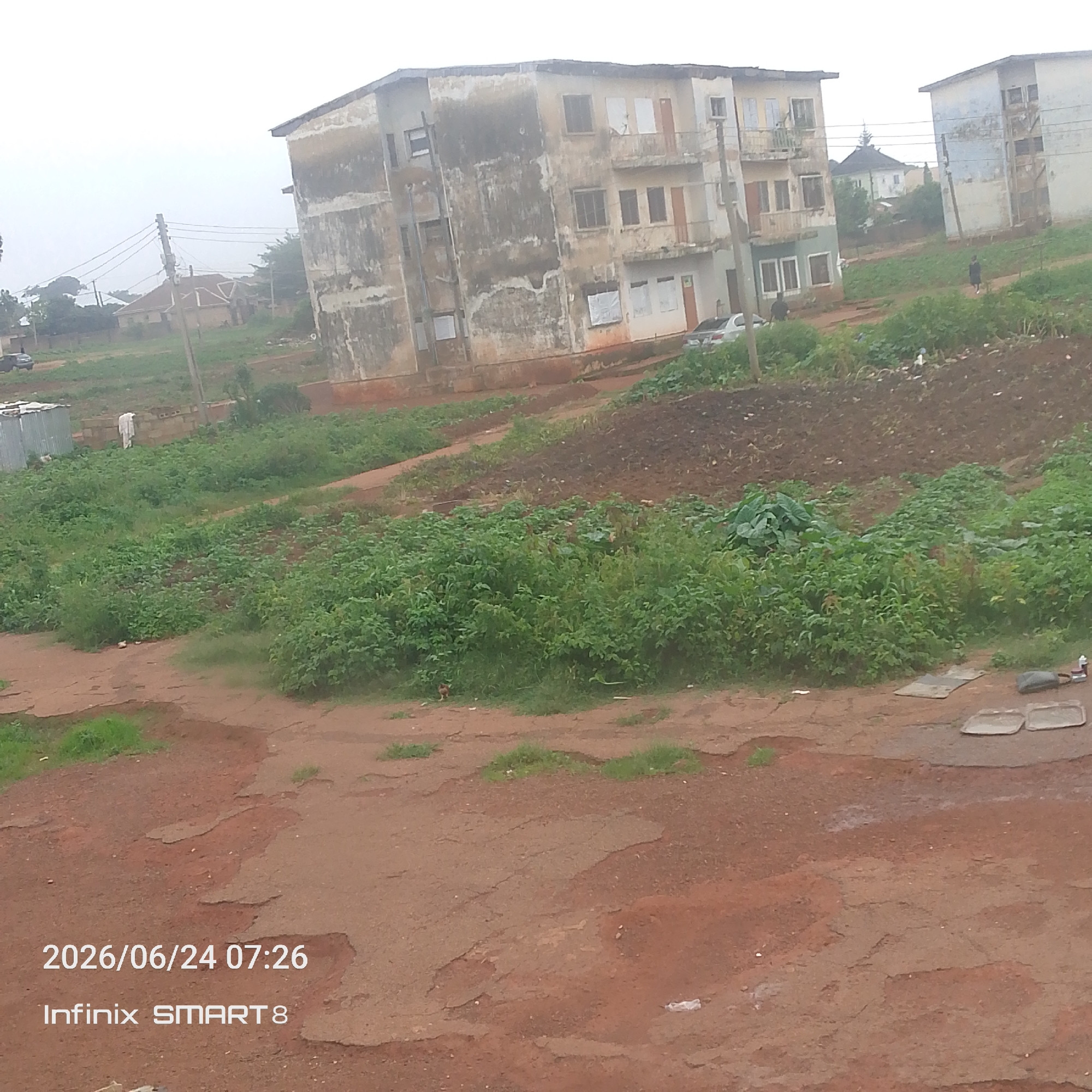 The image depicts an outdoor scene of a residential area, possibly in a developing country. In the foreground, a rough, reddish-brown dirt path occupies a significant portion of the frame, showing signs of wear and tear with cracks and uneven surfaces. Patches of green grass are interspersed throughout the path.
Further into the mid-ground, there are areas of lush green vegetation, appearing to be crops or garden plots. Beyond this, a dilapidated, multi-story building stands prominently, its facade weathered and stained, showing signs of age and disrepair. The building has multiple windows, some boarded up, and balconies. To the right of this building, another similar structure is partially visible, also appearing aged.
In the background, more buildings and trees are discernible against a hazy, overcast sky, suggesting a cloudy or misty day. A utility pole with electrical wires crosses the scene. A few small objects, possibly discarded items, are scattered on the ground.
The image has a watermark at the bottom left corner displaying "2026/06/24 07:26" and "Infinix SMART 8". There are no clear indications of specific activities or events taking place, other than the general presence of a residential environment. The overall mood conveyed is one of a somewhat neglected, rural or semi-urban setting under a somber sky.