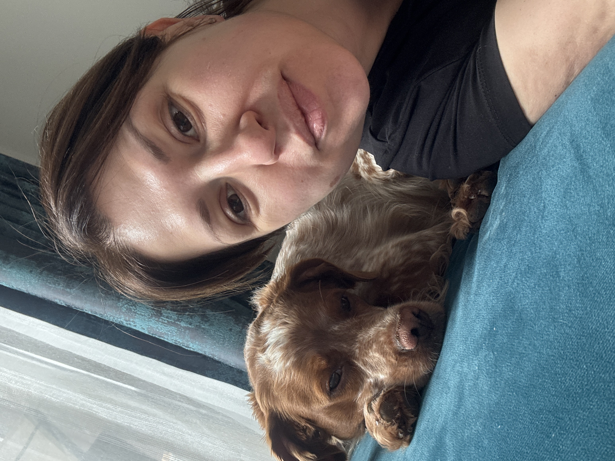 A woman lies next to a brown and white dog on a blue upholstered surface. The woman has brown hair and is wearing a black shirt. The scene appears to be indoors near a window with sheer white curtains. There is no indication of the location being in Sofia, Bulgaria.
