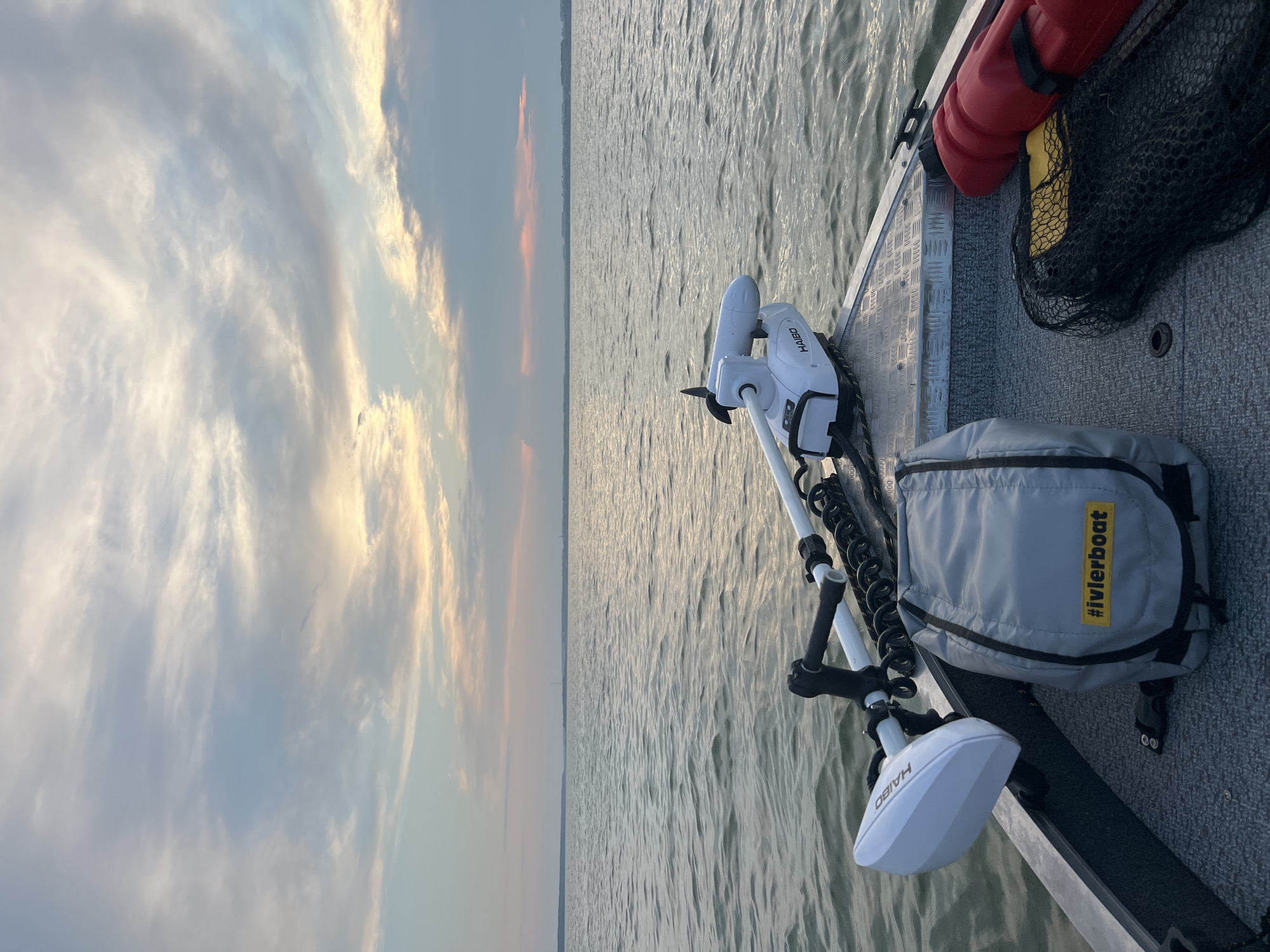 The image shows a view from the bow of a boat on a large body of water. The water is calm with small ripples. The sky above is filled with wispy clouds, with some areas of pink and orange suggesting a sunset. In the foreground, on the boat's deck, are several items: a white trolling motor with the brand "HAIBO" visible, a grey bag with the yellow hashtag "#ivlerboat" printed on it, a black fishing net, and a red fuel can. The boat appears to be made of aluminum with a gray carpeted deck. In the distance, a faint shoreline with some structures is visible. The location is specified as Novopichugovo, Russia. There are no people or animals visible in the image.