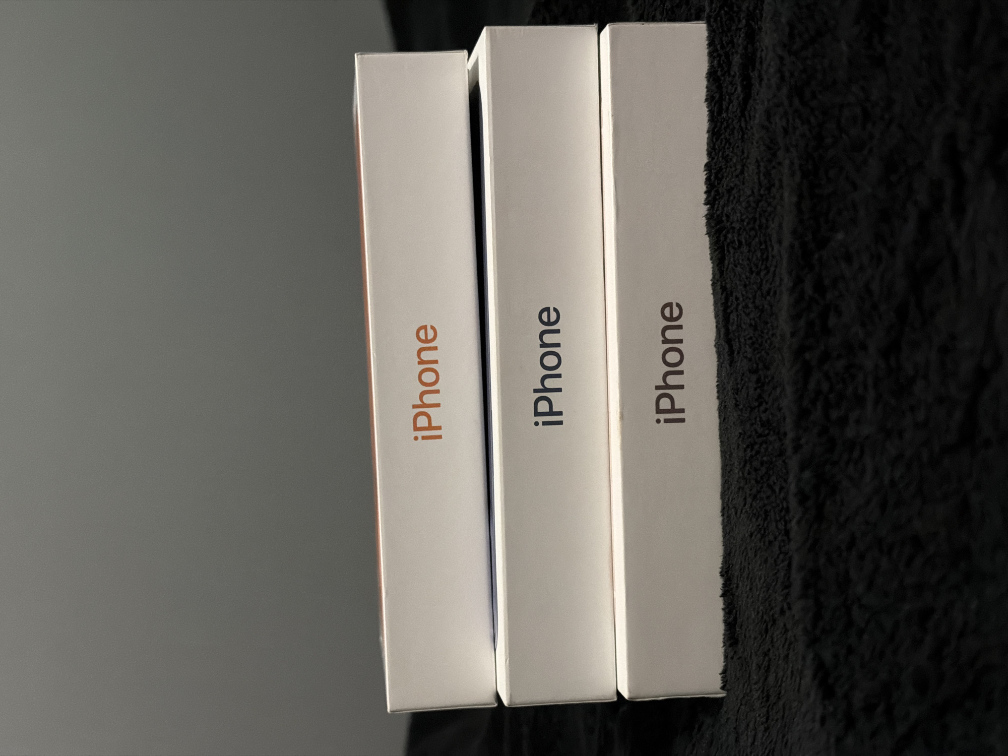 The image shows three iPhone boxes standing side-by-side, with the word "iPhone" printed vertically on each. The boxes are white. The word "iPhone" is in orange on the box to the left, and gray on the other two. The background is a gray wall on the left and a dark, textured surface on the right. The location in Varna, Bulgaria is unknown.