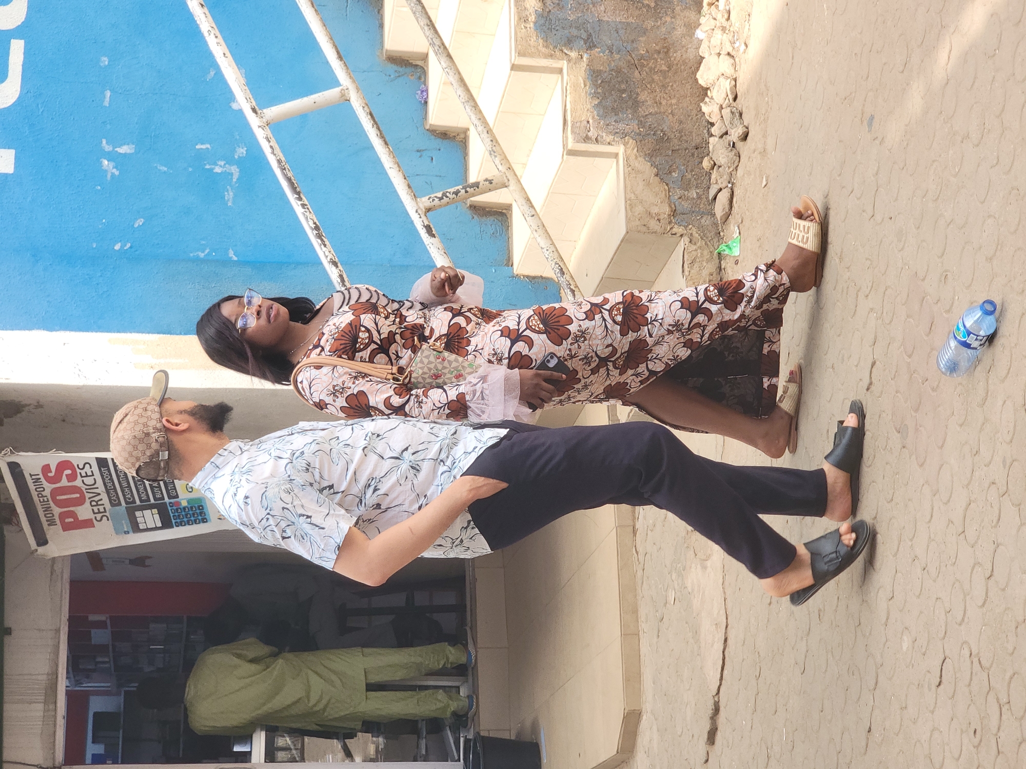 Here's a detailed description of the image:
**Scene Overview:**
The image appears to be taken on a sidewalk in what is identified as Jos, Nigeria. The scene features two people, a man and a woman, standing near a storefront. The setting seems to be a sunny day with outdoor lighting.
**People:**
* **Woman:** She is wearing a long, patterned dress with floral designs, along with sunglasses. She carries a small purse on her shoulder. She is fair-skinned. Her hair is black. She holds a cell phone.
* **Man:** He is wearing a patterned shirt, dark trousers, and black sandals. He has a beard and is wearing a baseball cap.
**Objects:**
* **POS Services Sign:** A sign for "MONIEPOINT POS SERVICES" is visible near the storefront, indicating financial services are offered. The sign details services such as cash deposit, cash withdrawal, and money transfer.
* **Building Exterior:** The building in the background has a blue wall with a metal railing and steps.
* **Water Bottle:** A water bottle is on the ground near the edge of the sidewalk.
* **Sidewalk:** The sidewalk is a textured, light-colored pavement.
**Environment:**
* **Location:** Identified as Jos, Nigeria.
* **Setting:** Appears to be an urban or commercial area.
* **Weather:** Sunny day with clear lighting.
**Actions and Interactions:**
* The woman and man are standing next to each other, possibly talking. Their body language appears casual.
* The man's hand is tucked in his pocket.
* The woman holds a cell phone in her hand.