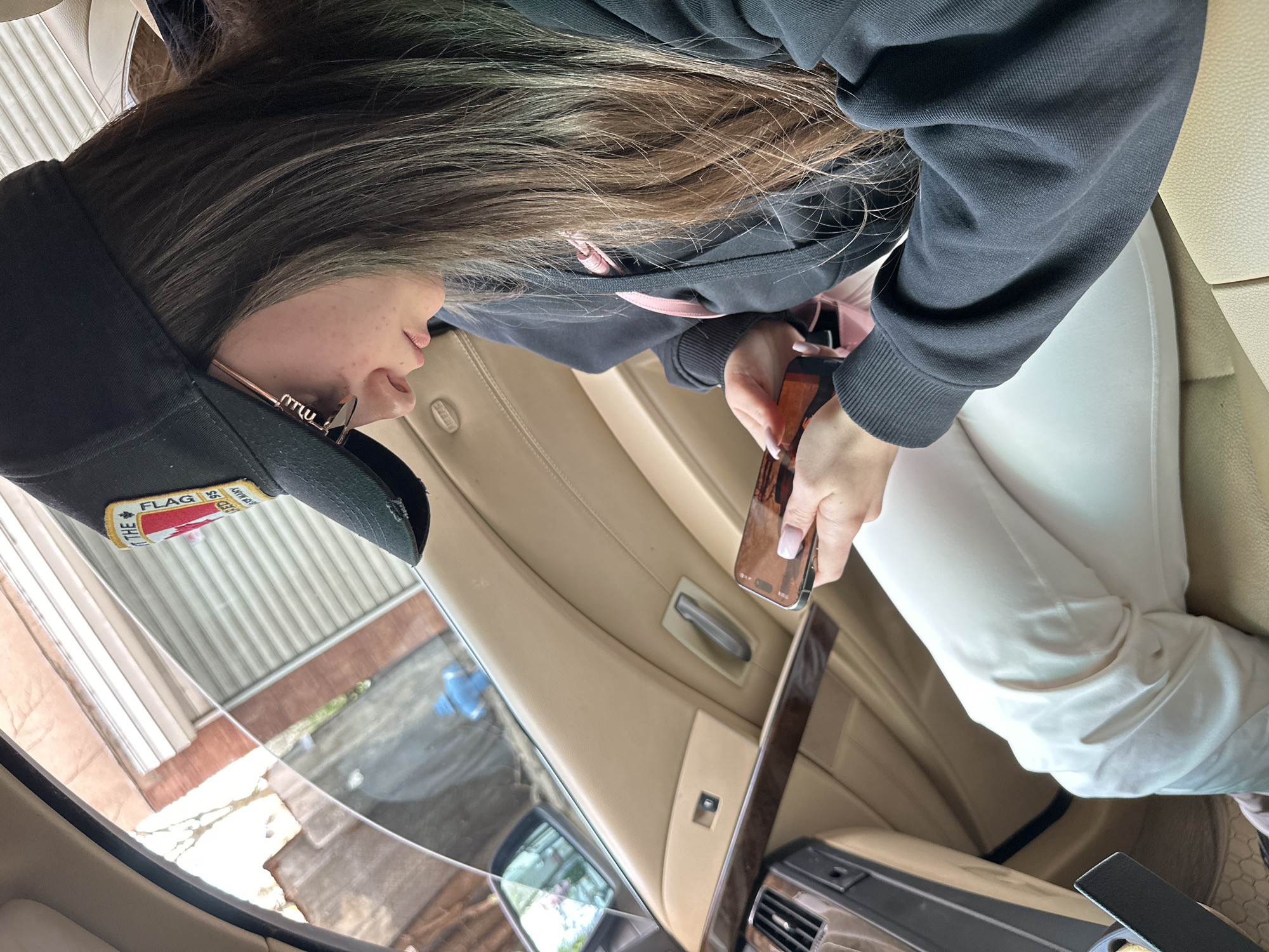 A woman sits in the passenger seat of a vehicle, holding a smartphone. She is wearing a black hoodie with pink accents, white pants, and a black baseball cap with a "The Flag" patch. The car's interior is beige leather and wood trim. The exterior visible through the car window shows a residential setting, suggesting the location could be in Plovdiv, Bulgaria.