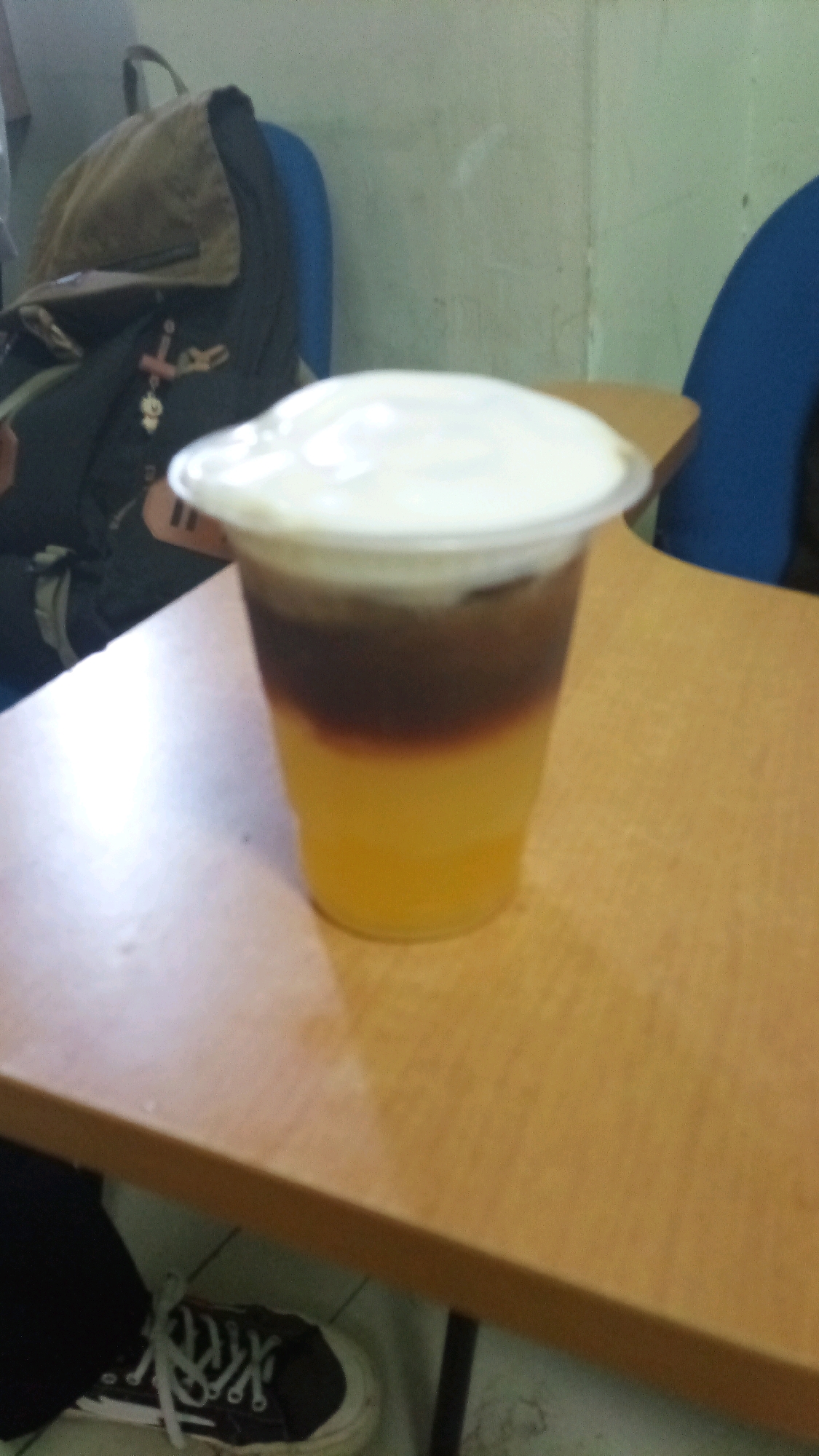 A close-up, eye-level, medium shot captures a layered beverage in a clear plastic cup with a white lid. The drink has three distinct layers: a bright yellow liquid at the bottom, a dark brown liquid in the middle, and a thick white foam topping. The cup sits on a light brown wooden table. In the background, a portion of a dark backpack with a brown flap is visible. To the right of the backpack, a dark blue chair back is partially seen. The wall behind the backpack appears to be light green or beige. The floor beneath the table shows a black shoe with white laces. The scene is indoors, likely in a casual setting such as a classroom or cafe in Lenek, Indonesia.