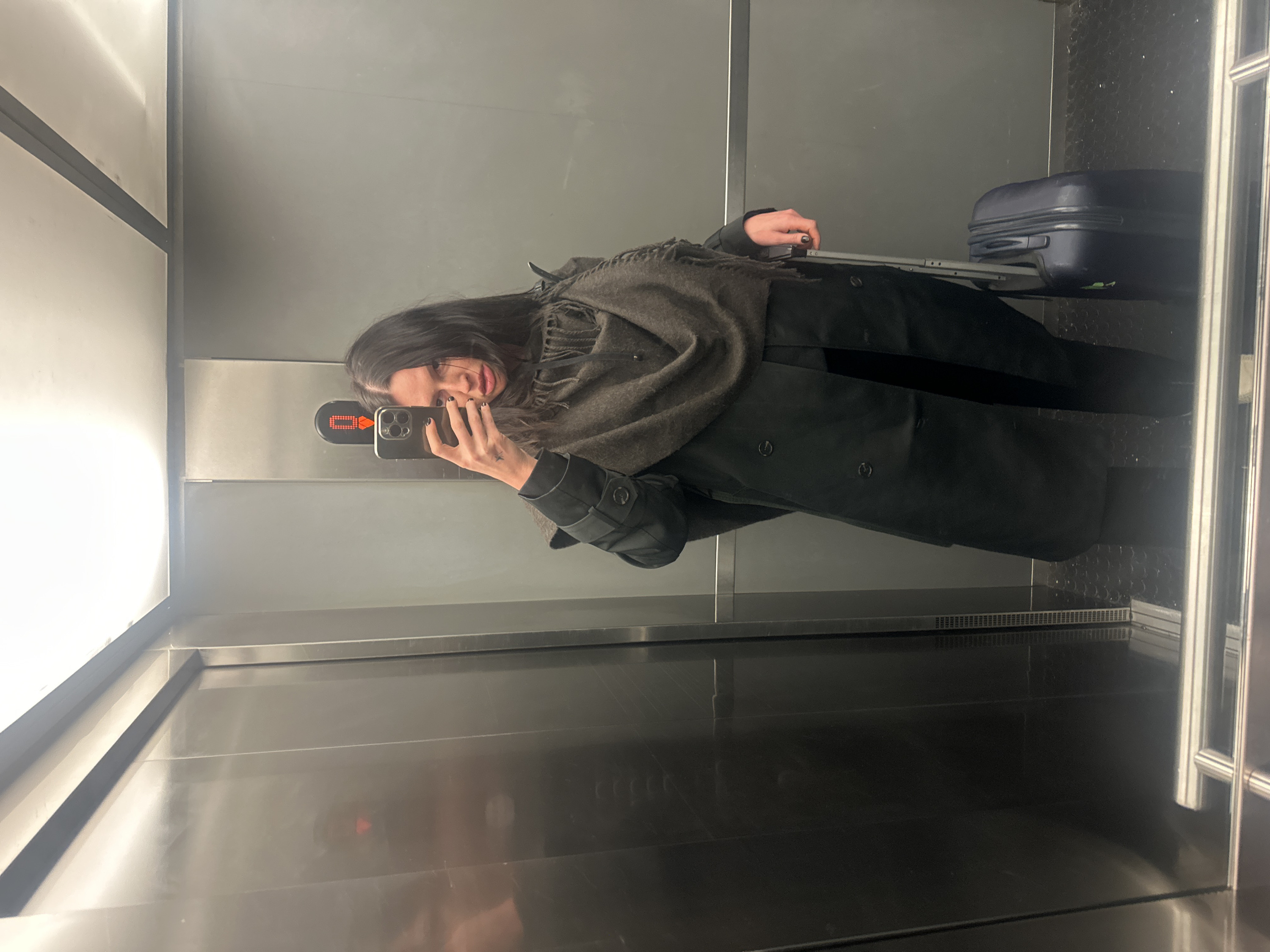 Inside an elevator, a person wearing a dark coat and gray scarf takes a selfie with a smartphone. The elevator walls are a reflective metallic gray. An illuminated digital display indicates the floor. A suitcase rests near the back wall. The floor of the elevator has a dark, glossy finish, reflecting the interior elements.