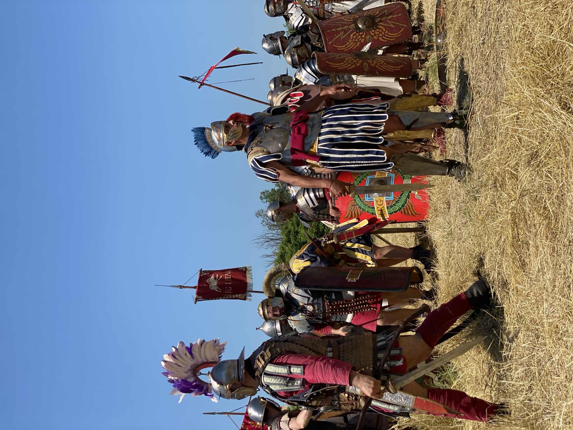 The image depicts a historical reenactment scene featuring a group of individuals dressed as Roman soldiers, assembled outdoors under a clear blue sky. The setting is a dry, golden-brown grassy field, with sparse green foliage visible in the distant background. The sun is bright, casting strong shadows.
In the foreground on the left, a man in full Roman legionary attire stands, facing towards the right. He wears a silver-colored helmet adorned with a large, elaborate purple and white feather plume. His body is covered by scale or segmented armor over a red tunic, and he has red leg wraps or trousers with black boots. He holds a gladius (short sword) pointed downwards in his right hand.
To his right and slightly behind him, another figure, partially visible, wears a silver helmet. Further right, a man with a bronze-colored helmet and what appears to be a horn-like crest stands, wearing armor over a red tunic and holding a dark red rectangular scutum (shield).
Dominating the mid-ground center is a prominent figure, likely an officer, facing slightly left. He wears a gleaming silver helmet with a distinctive blue feather plume. His torso is protected by a metallic chest plate (lorica musculata) over a white tunic with vertical blue stripes on its pteruges (skirt-like strips). A red sash is tied around his waist. He holds a short sword or dagger at his left hip.
To the right of this central figure, several other individuals in varied Roman military dress are visible, including helmets, armor, and shields. One person carries an upright spear with a simple red banner. Another holds a red scutum decorated with a golden wreath surrounding what appears to be a lion or eagle head, and the letters "LEG".
In the background, on the left side, a red vexillum (standard) is visible on a pole, featuring a white animal emblem (possibly a boar or horse) and the inscription "LEG I ITAL".
The individuals are standing, posing, or awaiting instructions, indicating a staged event rather than active combat. This scene takes place in Gigen, Bulgaria.