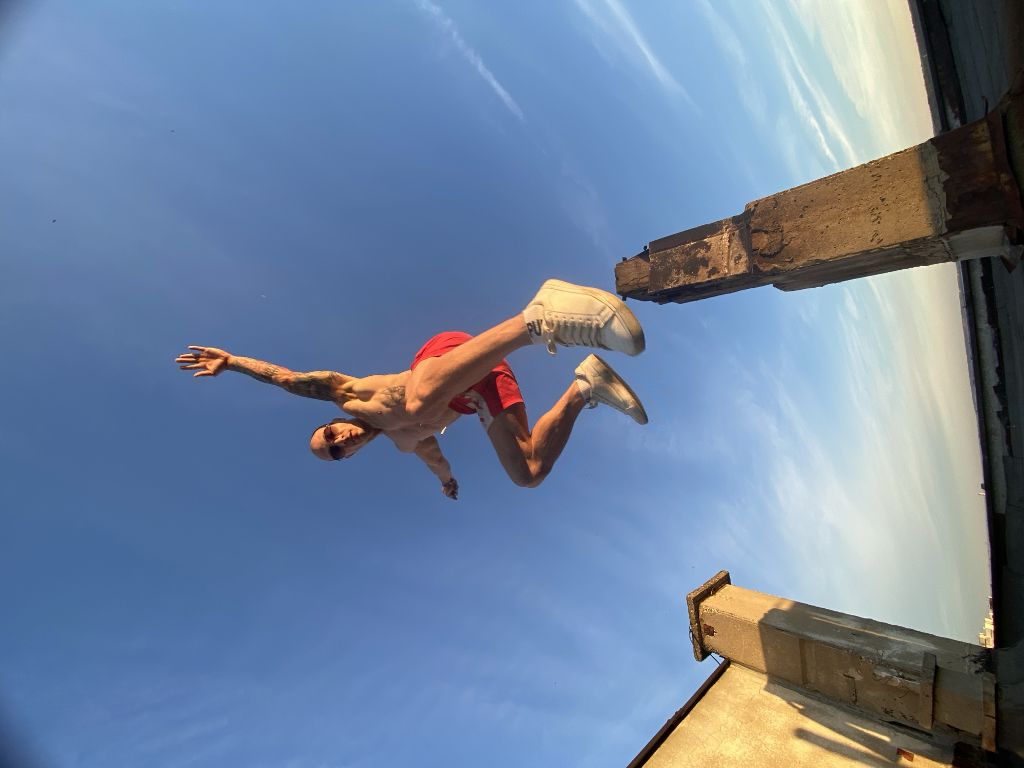 A man is captured mid-air from a low-angle perspective against a blue sky with wispy clouds. He is wearing red shorts, white sneakers, and sunglasses. His arms are extended, and his body is angled as if in a jumping or flying pose. The foreground consists of the corner of a weathered concrete structure and parts of a building. The location is Pleven, Bulgaria.