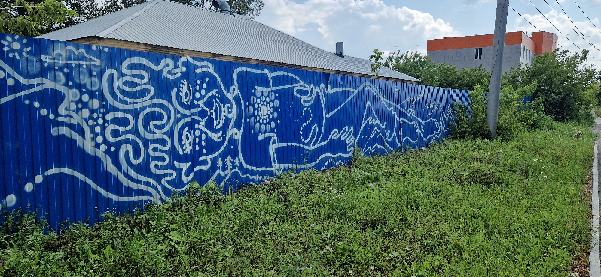The image depicts a long, blue corrugated metal fence with white graffiti art. The graffiti is abstract and features swirling lines, dots, and shapes that resemble natural elements like mountains and clouds. The fence is located in a city setting, as indicated by the presence of a building with orange and gray siding in the background, as well as utility poles and wires. The fence is bordered by a patch of overgrown green grass and weeds, suggesting it might be in a less maintained area or an undeveloped lot. The sky is visible above, with some clouds present. The overall scene is mundane, showcasing urban decay and artistic expression through graffiti.