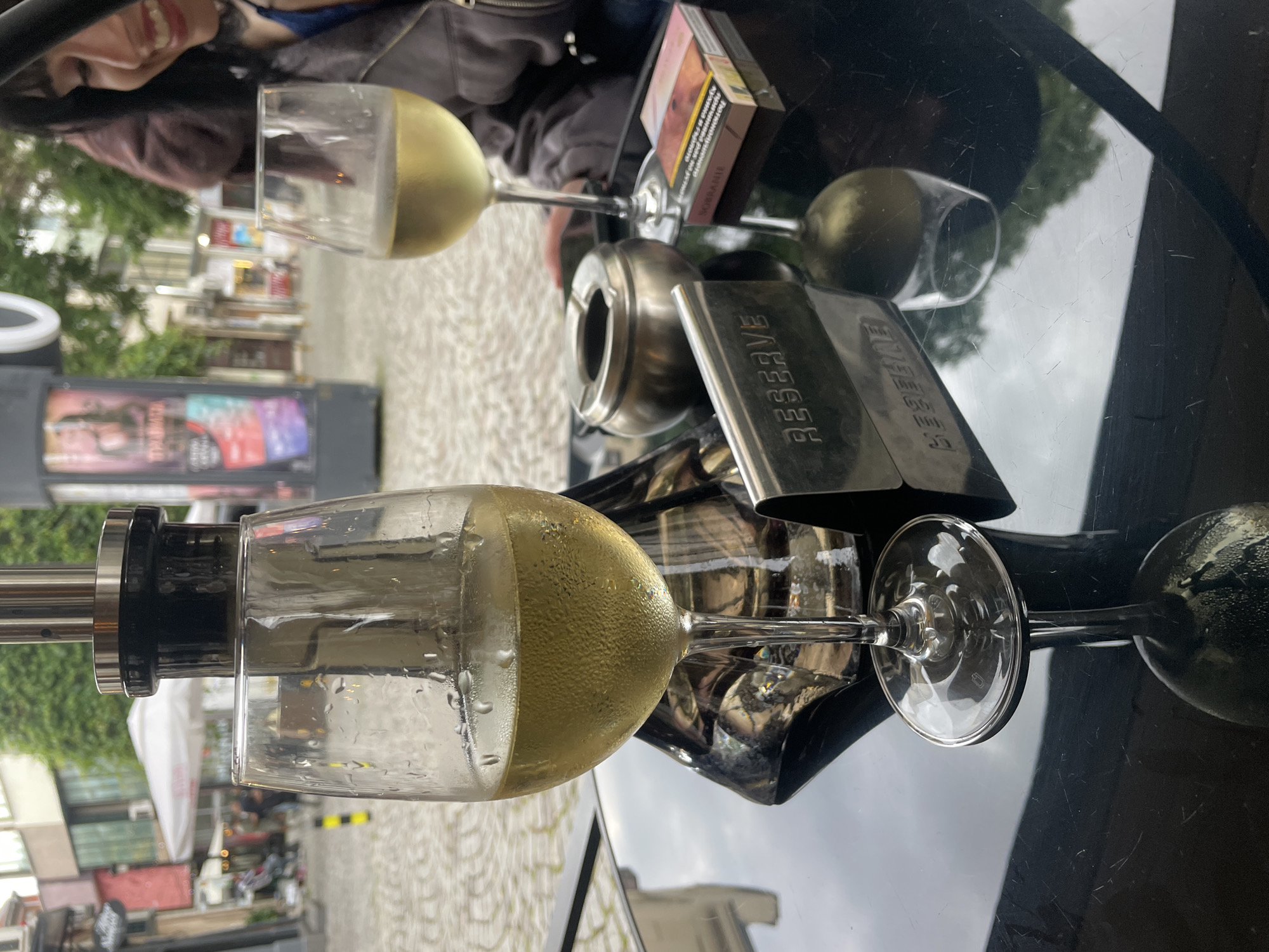 A medium shot captures a table setting at an outdoor cafe. In the foreground, a glass of white wine, condensation visible on the glass, sits on a black reflective table. To the right of the wine glass, another partially visible glass of white wine is present. An ashtray, a cigarette pack, and a metal "RESERVE" sign are also on the table. In the background, a woman with dark hair is partially visible, smiling. The cafe appears to be situated on a cobblestone street with buildings and trees in the distance. The overall lighting suggests an overcast day.