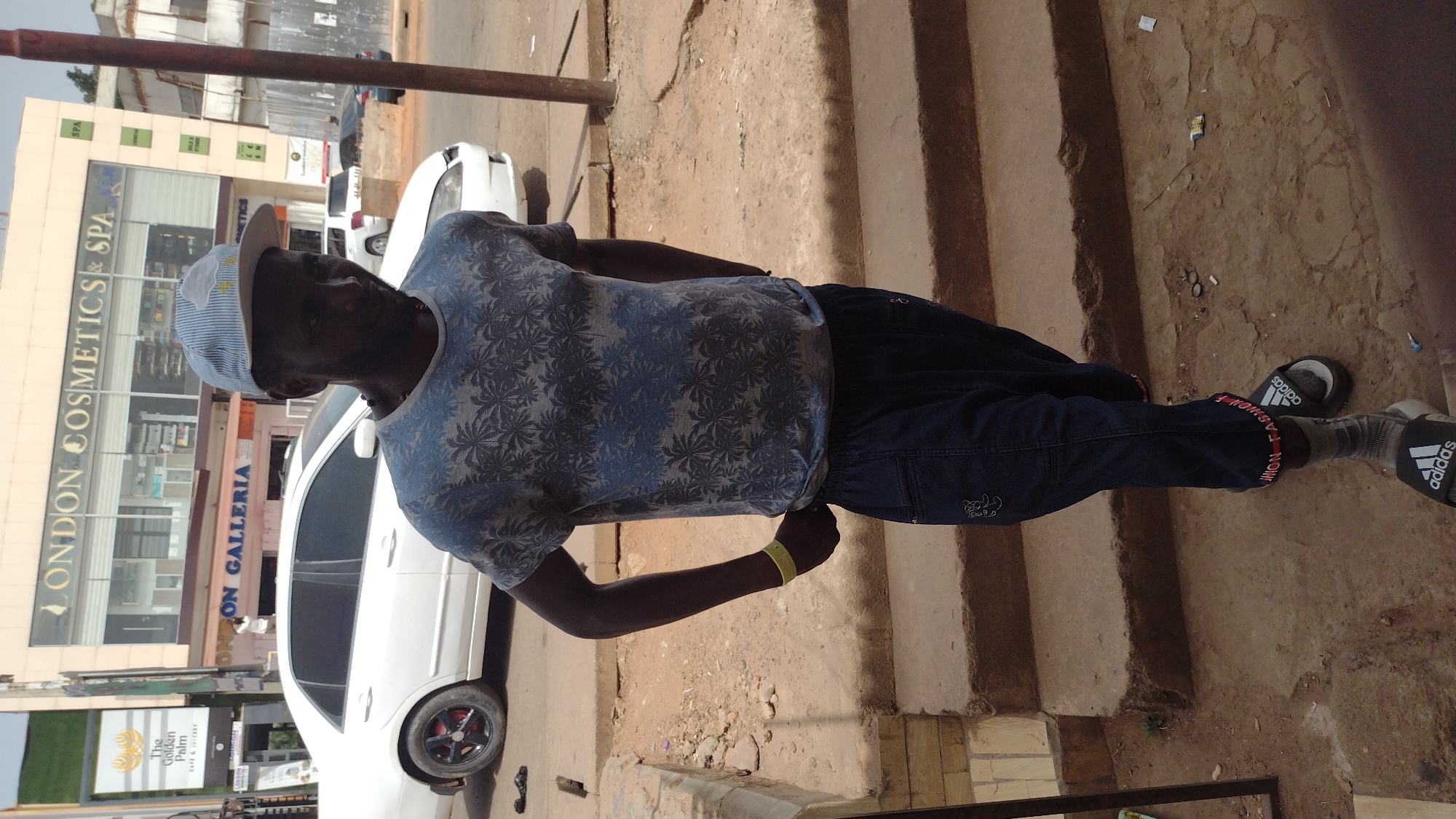 A medium, eye-level, outdoor shot captures a man in Jos, Nigeria, standing on a set of concrete steps next to a street. He is wearing a light blue, palm-tree-patterned t-shirt, dark-blue pants, a blue-striped bucket hat, grey socks, and black Adidas sandals with red and white lettering. He has a yellow wristband on his left wrist. He stands beside a white car parked on the side of the road.
In the background, a building prominently displays the signs "LONDON COSMETICS & SPA" and "LONDON GALLERIA." To the left, another sign reads "The Golden Palm CAFE & JUICERY." Other parked cars are visible along the street. The sky appears pale, suggesting daylight. The ground around the steps is dirt and debris-strewn. A small green plant grows beside the steps.