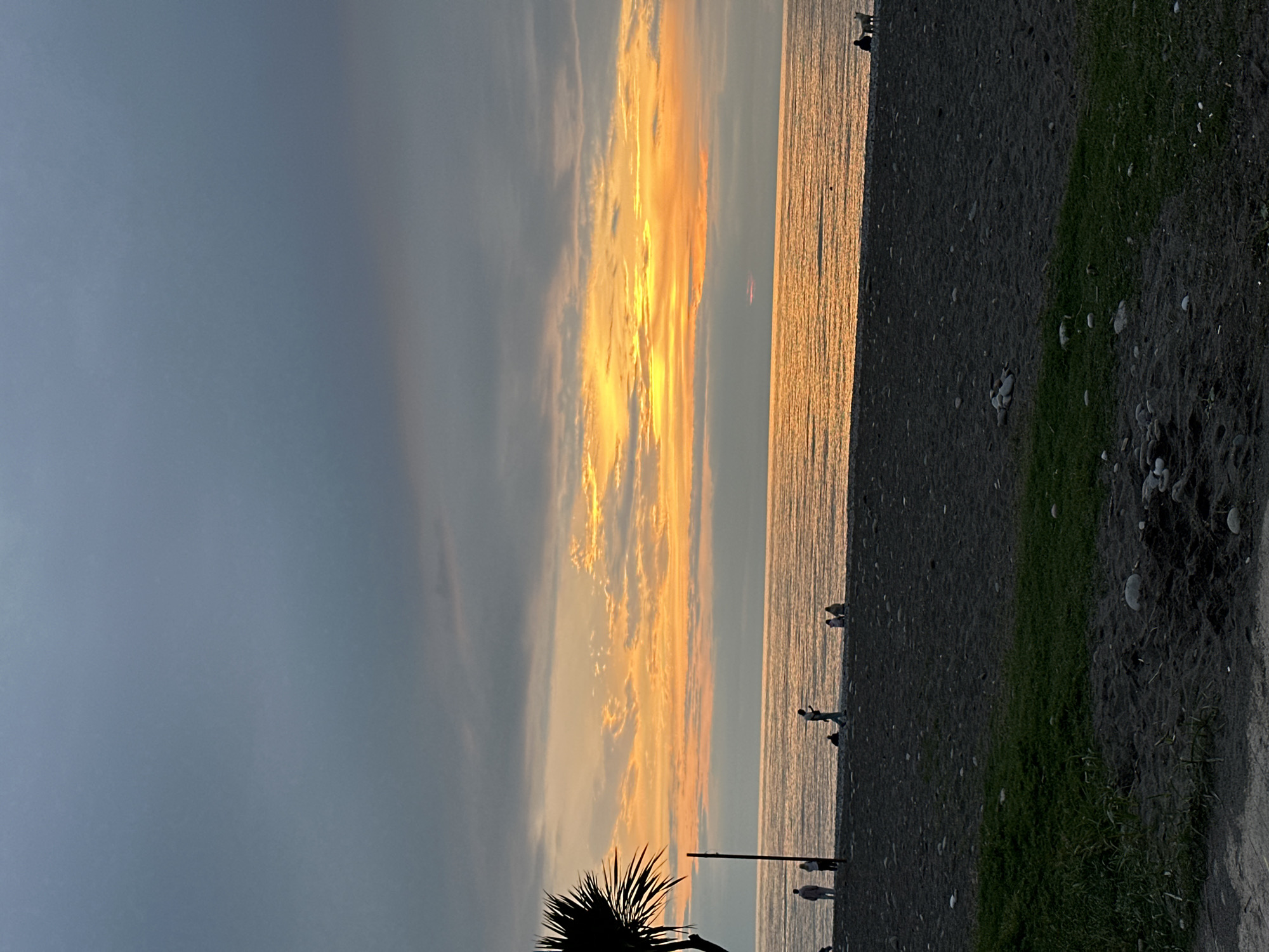 The image depicts a sunset view of the beach in Batumi, Georgia. The sky is a mix of dark gray and orange hues, reflecting on the water. Several people are walking along the dark sandy beach. Vegetation, including grass and a palm tree, is visible along the edge of the beach.