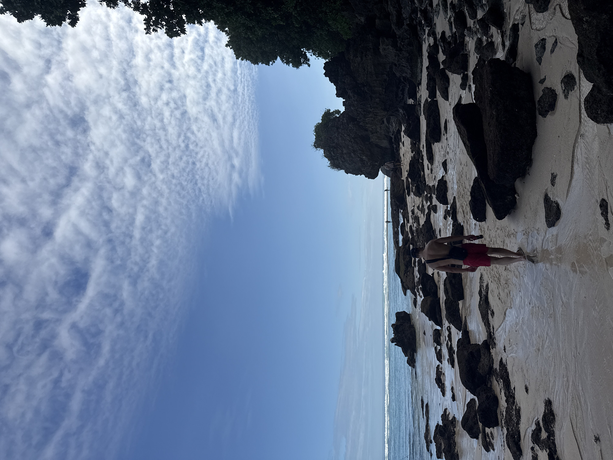The image shows a beach scene in Kuta Selatan, Indonesia. A man in red shorts walks along the shoreline amongst rocks and sand. The sky is blue with scattered clouds. The ocean is visible in the background. Vegetation is present at the top of the image.