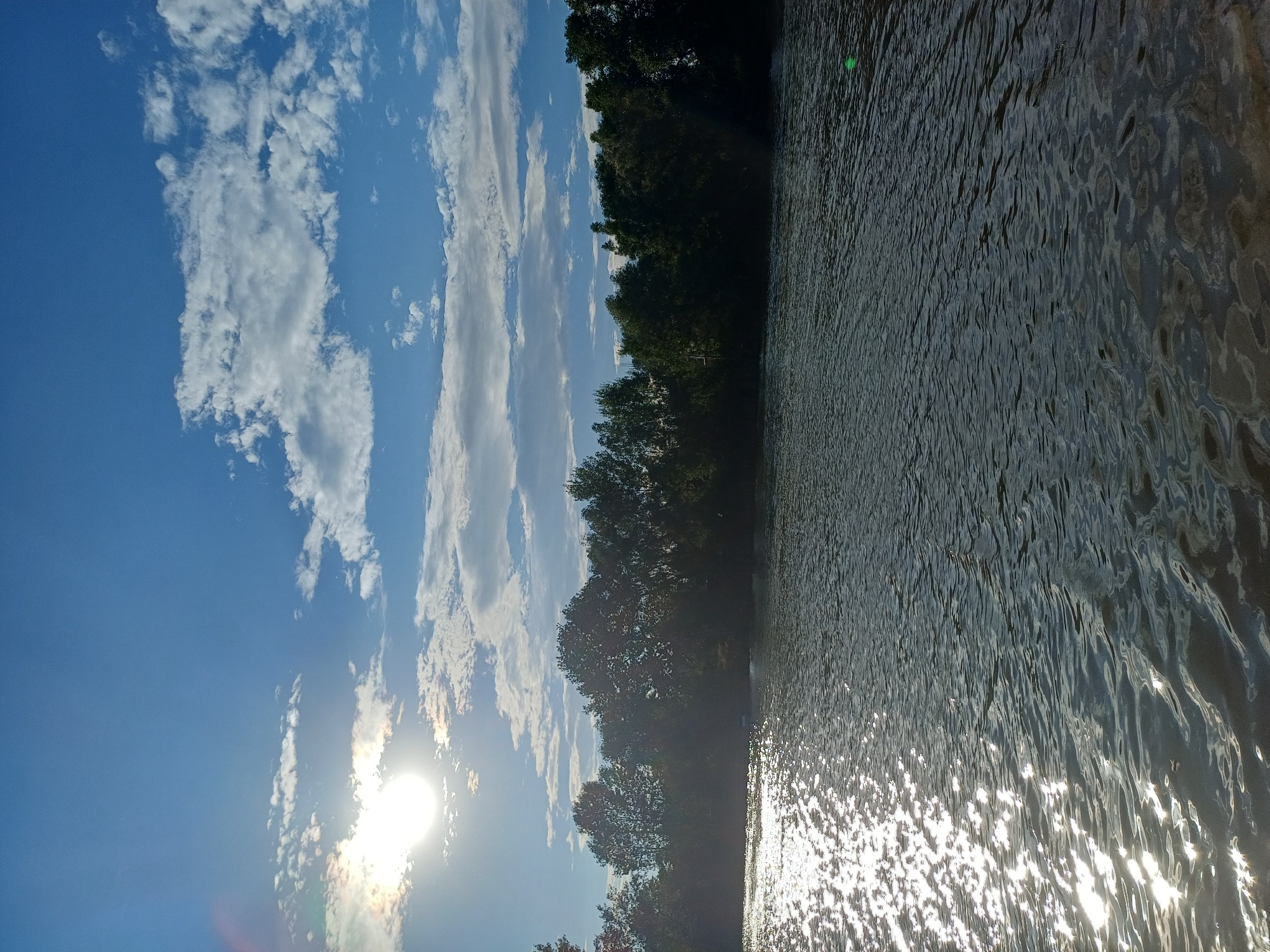 The image depicts a body of water, likely a river, with sunlight reflecting off its surface, creating a shimmering effect. The water is dark and rippled, suggesting a gentle current or breeze. Along the far bank of the water, a dense line of trees forms a natural border, their foliage casting shadows. Above the trees, a bright sun shines through a blue sky dotted with scattered white clouds. The scene appears to be a natural landscape, possibly a park or a riverside area within the city of Sulymivka, Ukraine, given the context provided. There are no people or specific man-made structures prominently visible in this serene natural setting.