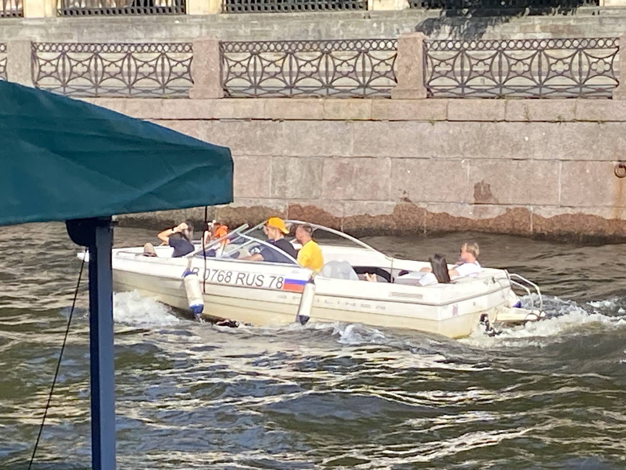 A white speed boat with four occupants is moving on the water. The boat is registered with the number "0768 RUS 78" and features a Russian flag decal. In the foreground, a green awning covers part of the view. The background shows a stone embankment with an ornate metal railing, characteristic of Saint Petersburg's architectural style. One person in the boat is wearing a yellow cap. The water is choppy, indicating the boat is in motion.