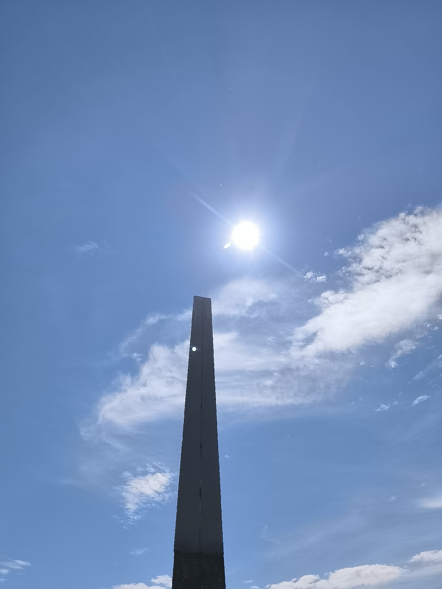 The image shows a tall, slender, dark gray obelisk against a bright blue sky with scattered white clouds and the sun. The obelisk is positioned in the lower half of the frame, extending upwards. A bright lens flare from the sun is visible near the top of the obelisk. The obelisk appears to be made of a smooth, matte material and has sharp, straight edges. The sky is clear and vast, with wispy clouds suggesting fair weather. The overall impression is one of a monumental structure set against a natural backdrop. This obelisk is a landmark in Neringa, Lithuania, specifically in the town of Juodkrantė, marking the place where the German poet and writer Johann Sebastian von Dreyer was buried.