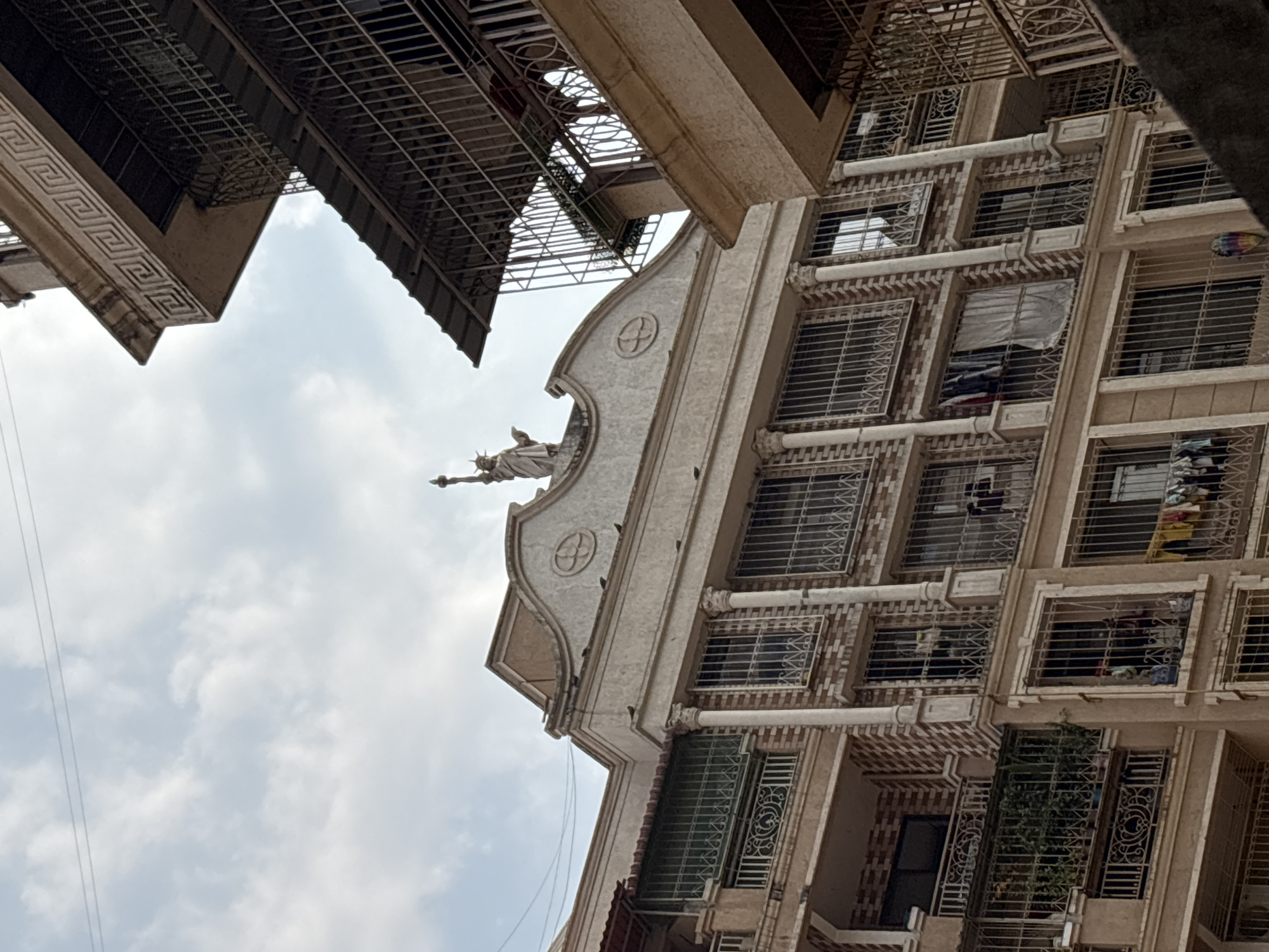 The image displays a multi-story residential building in Ambernath, India, under a cloudy, overcast sky. The central structure features a cream-colored facade with decorative elements, including vertical columns and sections of brick-patterned tiling. A prominent pediment-like structure crowns the building, upon which a miniature replica of the Statue of Liberty stands, holding a torch aloft in its right hand and a tablet in its left. Numerous windows are visible, all secured with intricate metal grates. Several balconies feature metal railings; on multiple balconies and windows, clothes are hanging to dry. Small birds, likely pigeons, are perched on the ledges and the pediment of the central building. To the right, a section of another building, possibly part of the same complex, features balconies with ornate metalwork and a cornice with a Greek key pattern. Electrical wires are discernible against the sky in the upper left quadrant. No human figures are present.