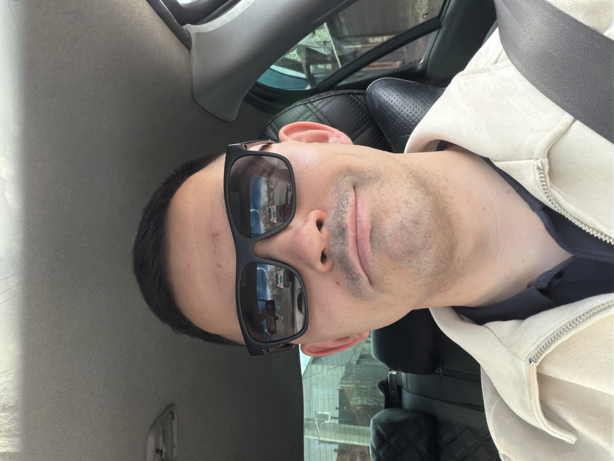 A close-up shot captures a man from the shoulders up, seemingly within the interior of a vehicle. He is seen with his head slightly tilted, wearing black rectangular sunglasses that obscure his eyes but clearly reflect an outdoor scene. The reflection in the sunglasses reveals a white car on what appears to be a street, alongside blurred buildings or other urban structures. The man has short, dark hair and visible stubble on his chin and jawline.
He is dressed in an off-white, perhaps cream-colored, hooded jacket with a silver zipper, worn over a dark blue undershirt. A grey seatbelt diagonally crosses his chest, indicating he is either a driver or passenger. The car's interior elements are visible around him: a dark, possibly leather, car seat with a diamond-stitched pattern to his right, and a grey headliner above and to his left. A car window is partially visible in the upper right, showing indistinct reflections of the outside environment.
The lighting suggests it is daytime and bright, likely sunny, given the use of sunglasses. The overall impression is of a casual moment captured inside a car. There is no visible text in the image. The location context for this media file is Izhevsk, Russia.