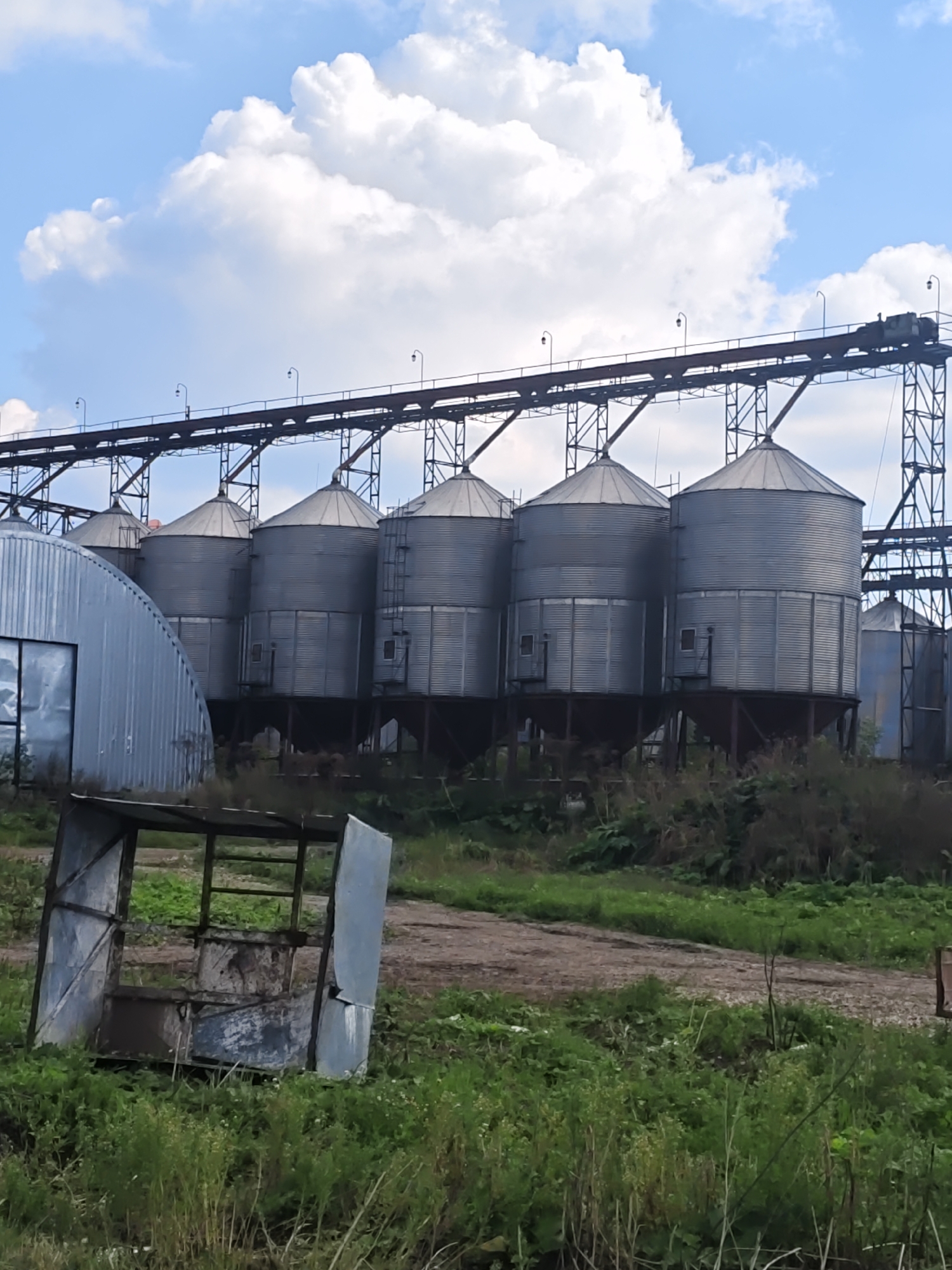 The image shows a row of industrial grain silos against a cloudy blue sky. The silos are tall, cylindrical structures made of corrugated metal, with conical roofs. They are supported by a network of metal beams and a conveyor belt system runs along the top. To the left of the silos is a large, curved metal building. In the foreground, there is a dilapidated metal structure, possibly a shelter or a piece of equipment, resting amidst overgrown green grass and vegetation. The ground appears to be a dirt path or area. The overall scene suggests an agricultural or industrial facility. The location is identified as Platoschino, Russia. There are no people visible in the image.