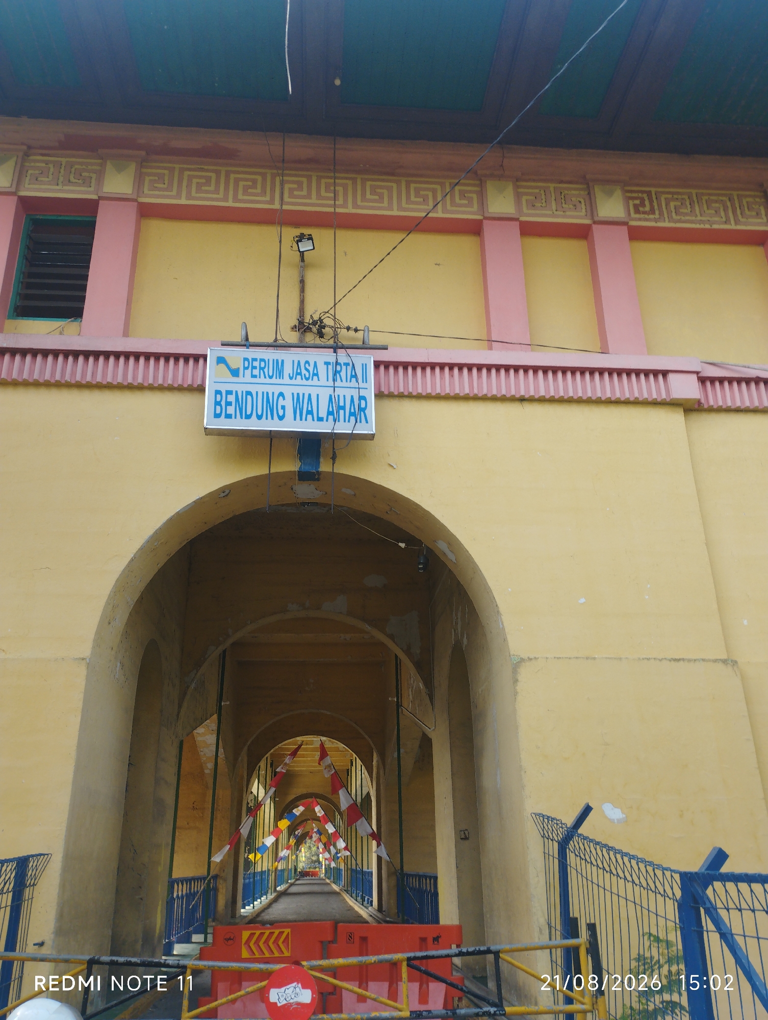 The image shows an arched structure painted yellow with pink accents, located in Karawang, Indonesia. A blue and white sign above the arch reads "PERUM JASA TIRTA II BENDUNG WALAHAR". Inside the archway, a pathway is visible, lined with red and white striped flags and blue railings. There are also red barriers and yellow chevron markings on the ground, suggesting a temporary closure or construction. The scene appears to be outdoors, with a green ceiling visible at the top of the frame. The overall impression is one of infrastructure, possibly related to water management or a dam.