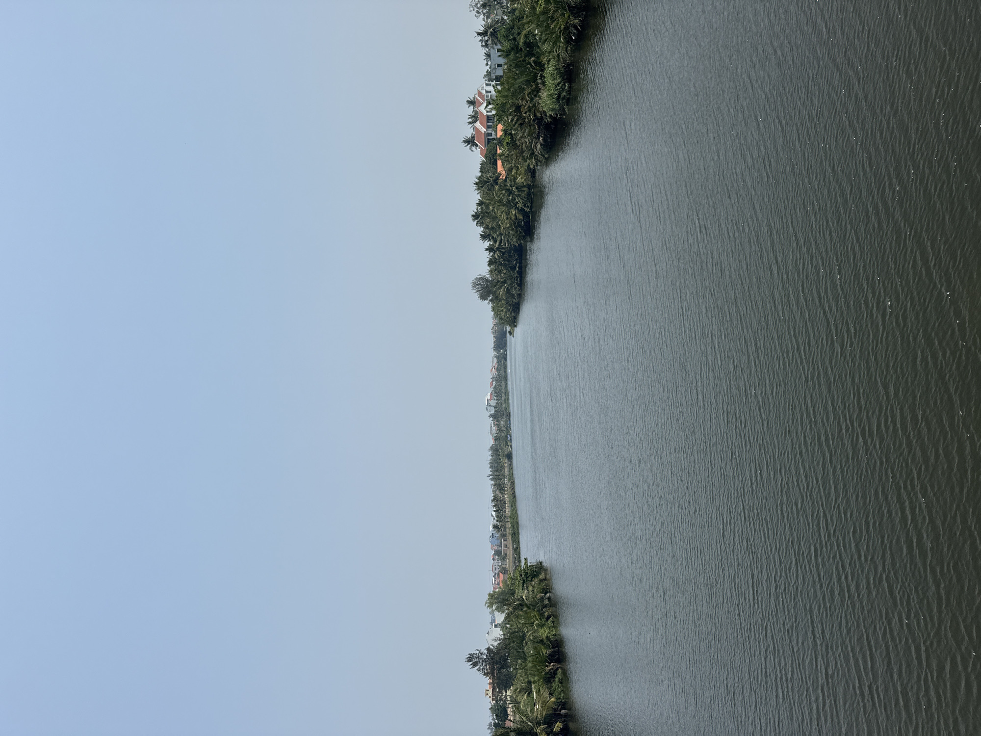 The image shows a long, dark body of water, likely a river or canal, running vertically through the frame. Along both banks are dense rows of green trees and other vegetation. On the right bank, there are a few buildings with red roofs visible among the trees. The left side of the image is predominantly a pale blue sky. The image is taken from a high vantage point. Based on the vegetation, architecture, and overall environment, the location is likely a river or canal in Đà Nẵng, Vietnam. No people or explicit actions are visible.