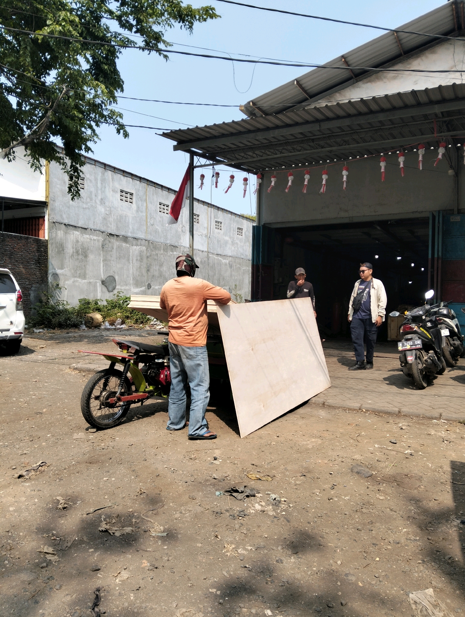 In Probolinggo, Indonesia, an outdoor scene unfolds in front of a workshop or warehouse with a corrugated metal roof supported by pillars. The ground is a mix of dirt and gravel, with some debris scattered about. To the left, a white car is parked. In the center of the image, a person wearing an orange shirt and blue jeans is standing next to a modified motorcycle, holding a large sheet of plywood. Behind this person, two other individuals are visible near the entrance of the workshop. One is wearing a dark shirt and a cap, and the other is wearing a light-colored jacket and a dark shirt. Several motorcycles are parked in front of the workshop. Red and white decorative paper lanterns hang in front of the workshop, along with a red and white Indonesian flag. The sky is clear and blue.