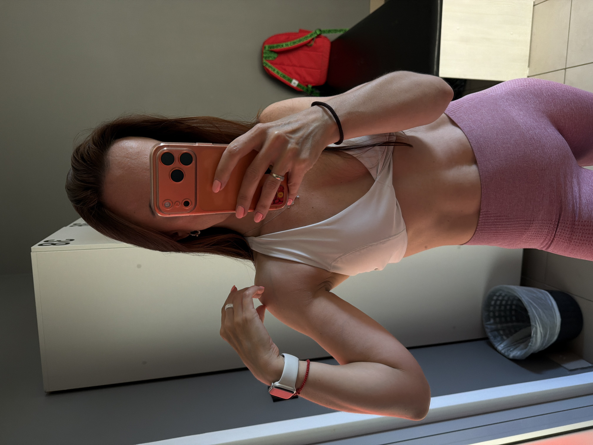 A woman is taking a mirror selfie in what appears to be a gym locker room. She is wearing a white sports bra and pink leggings, and her hair is brown and down. She is holding an orange smartphone in her right hand, which is reflecting the image back at the camera. On her left wrist, she is wearing a white smartwatch and a red bracelet. On her right hand, she is wearing a ring. In the background, there are lockers, a black bench, and a red backpack. There is also a trash can in the foreground. The lighting suggests it is daytime. The image does not contain any information that would allow for determination of its location in Birobidzhan, Russia, or any other specific city.