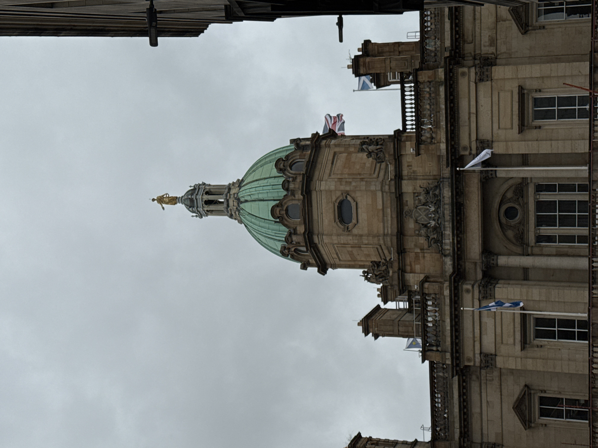 The image shows the dome of the former Edinburgh Sheriff Court building, located at 181 Causewayside, Edinburgh, United Kingdom. The building features a prominent green copper dome topped with a golden statue. Several flags are visible, including the Union Jack and the Scottish Saltire. The sky is overcast, suggesting a cloudy day. The architecture is classical with stone facades, columns, and decorative elements. The perspective is from below, looking up at the building.