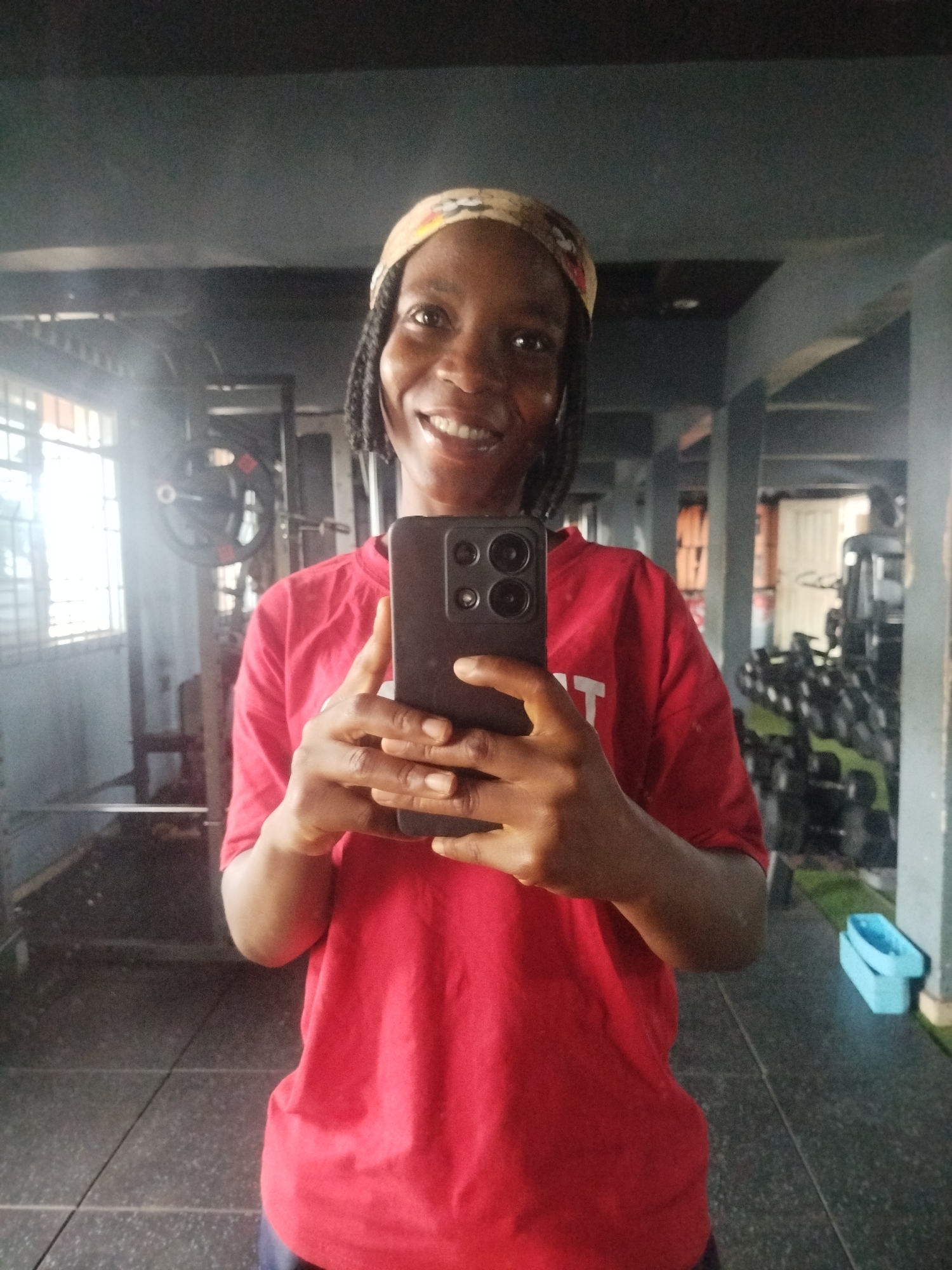 The image depicts a full-body mirror selfie of a young Black woman in a gym. She is wearing a red t-shirt and a patterned headband. She has dark braided hair and is smiling at the camera. She is holding a smartphone in her right hand, using it to take the selfie. The background of the image shows gym equipment, including weight racks and a window. A blue object is visible on the floor to the right of the woman. The woman's location is identified as Uselu, Nigeria, implying the gym is situated in that city.