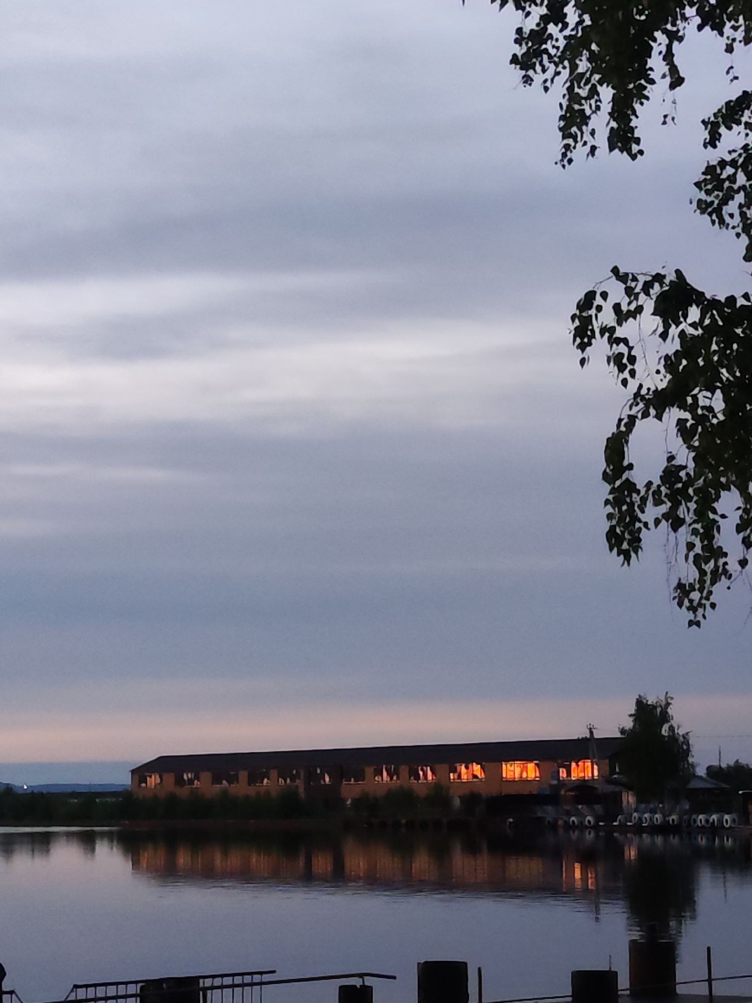 The image shows a long, one-story building on a body of water, likely a lake or wide river, in Chaikovsky, Russia. The building has several windows that are illuminated with warm light, suggesting activity inside. The building is reflected in the water, creating a symmetrical effect. In the foreground, there is a partially visible metal railing and some posts, suggesting a pier or dock. The sky is overcast with muted colors, indicating either early morning or late evening. A tree branch with leaves hangs in the upper right corner, adding a natural element to the scene. In the distance, a low-lying landmass is visible. The overall impression is one of a tranquil, lakeside setting at dusk or dawn. The repeated tires or similar round objects along the back of the building suggest its role as possibly a storage or service building for maritime activities.