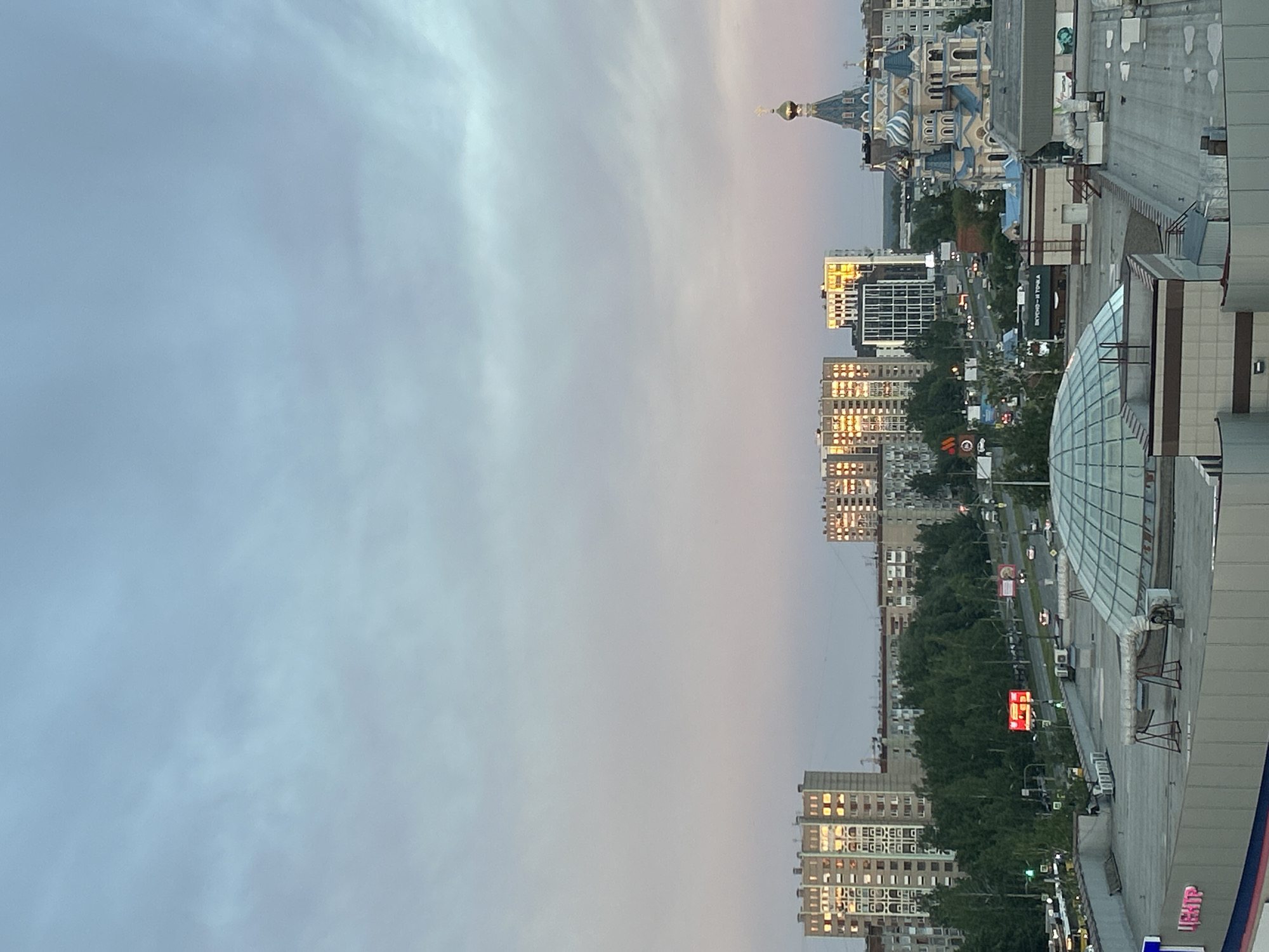 The image captures a cityscape in Izhevsk, Russia, during what appears to be either dawn or dusk, given the soft, muted light in the sky. The most prominent feature is the Mikhailovsky Cathedral, characterized by its blue domes and golden accents. The cityscape also features several high-rise buildings with reflective windows, indicating modern urban development. A shopping mall or commercial center is visible, identified by its glass dome and visible signage. The scene is observed from an elevated perspective, possibly from a higher building. A street runs through the cityscape, with some visible traffic and streetlights. The overall scene conveys an urban environment in transition between day and night.